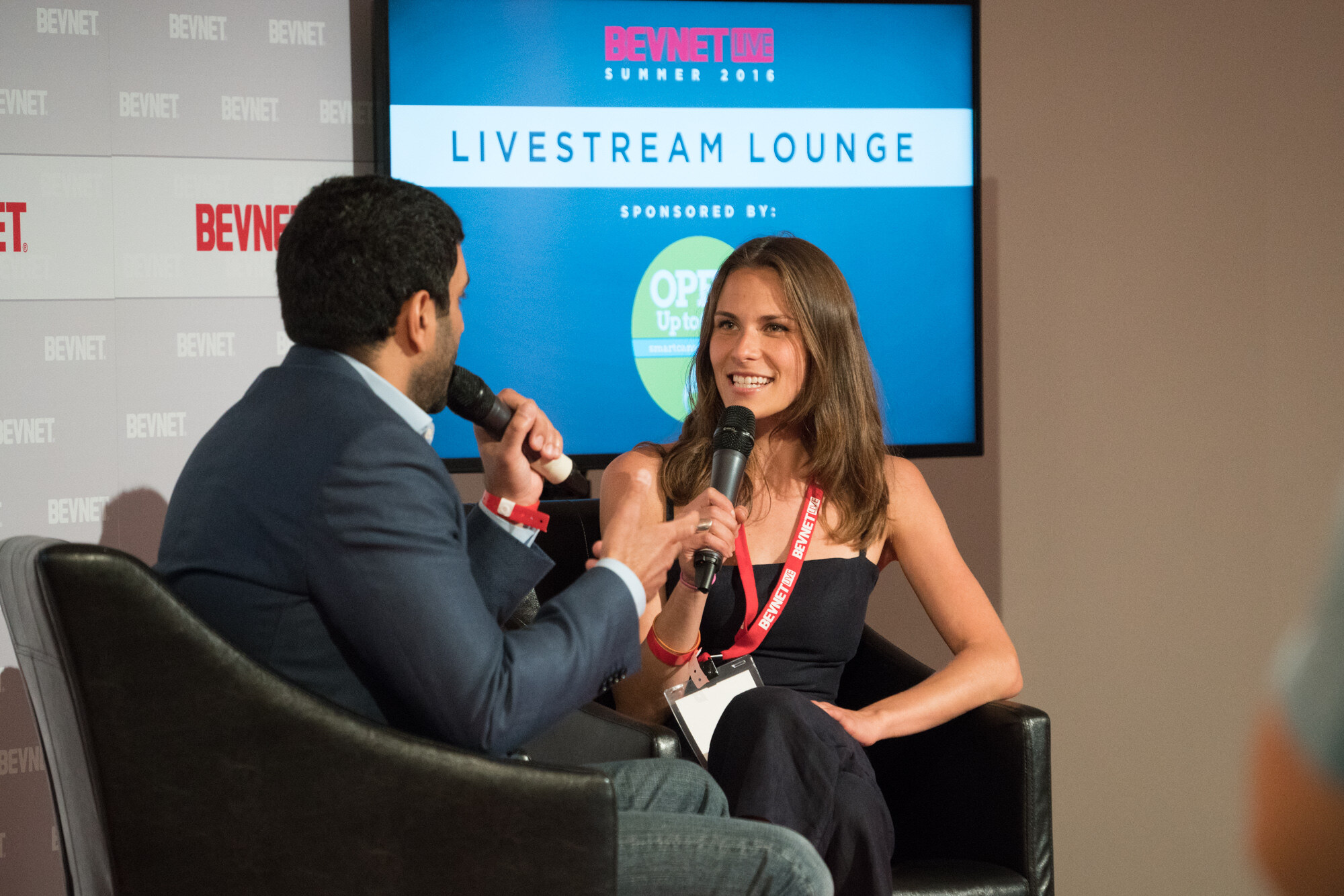 Woman with long brown hair and a BevNET Live lanyard holding a microphone and smiling toward an interviewer, with a screen reading Livestream Lounge, Summer 2016 behind her.