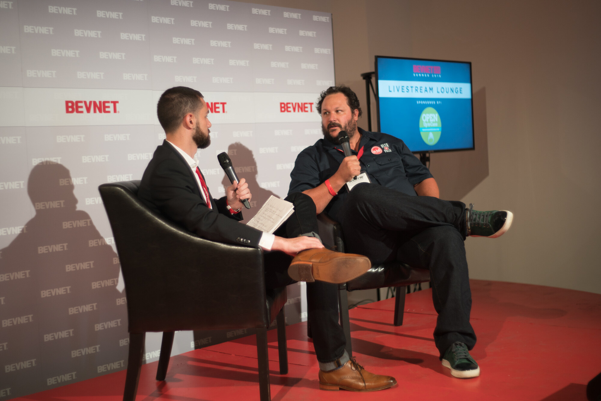 Two men in black leather chairs on a red platform, one in a dark suit with a notebook and one in a dark work shirt, with a Livestream Lounge screen sponsored by Open Up to Cans behind them.