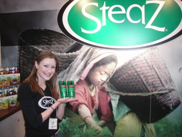 A smiling woman in a black Steaz shirt holds two green Steaz cans in front of a large Steaz sign and a photo of a person carrying a basket. Bottles of Steaz are stacked at left.