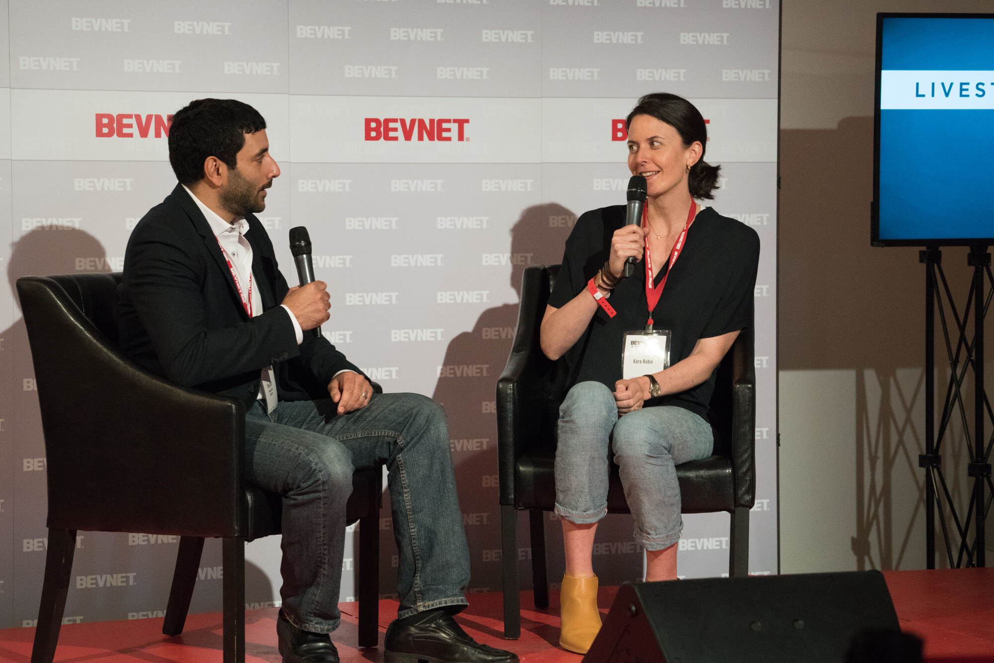 Two people in black leather chairs on a red platform in front of a BevNET step-and-repeat wall, the woman on the right holding a microphone and wearing a BevNET Live lanyard.