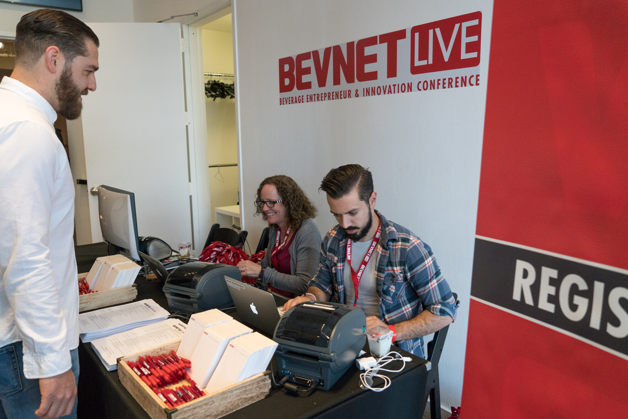 Two people seated at a black registration table with laptops, badge printers and stacks of badge holders, a bearded man in a white shirt at left and a BevNET Live sign on the wall behind.