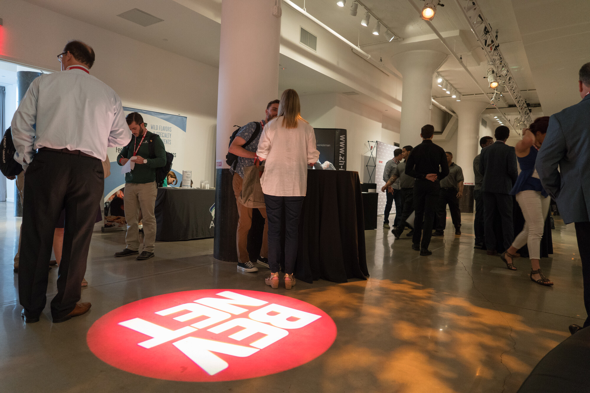 Low-angle view of a red BevNET projection on the concrete floor, with attendees standing at black-draped tables and exhibit banners behind.