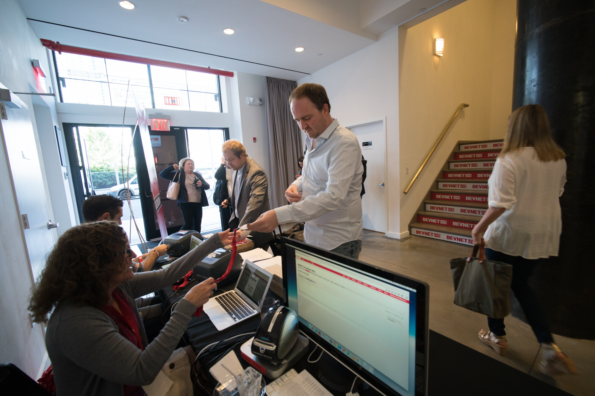 View over a registration computer of staff handing out lanyards to arriving guests, with a stairwell with BevNET step labels and the street doors behind.