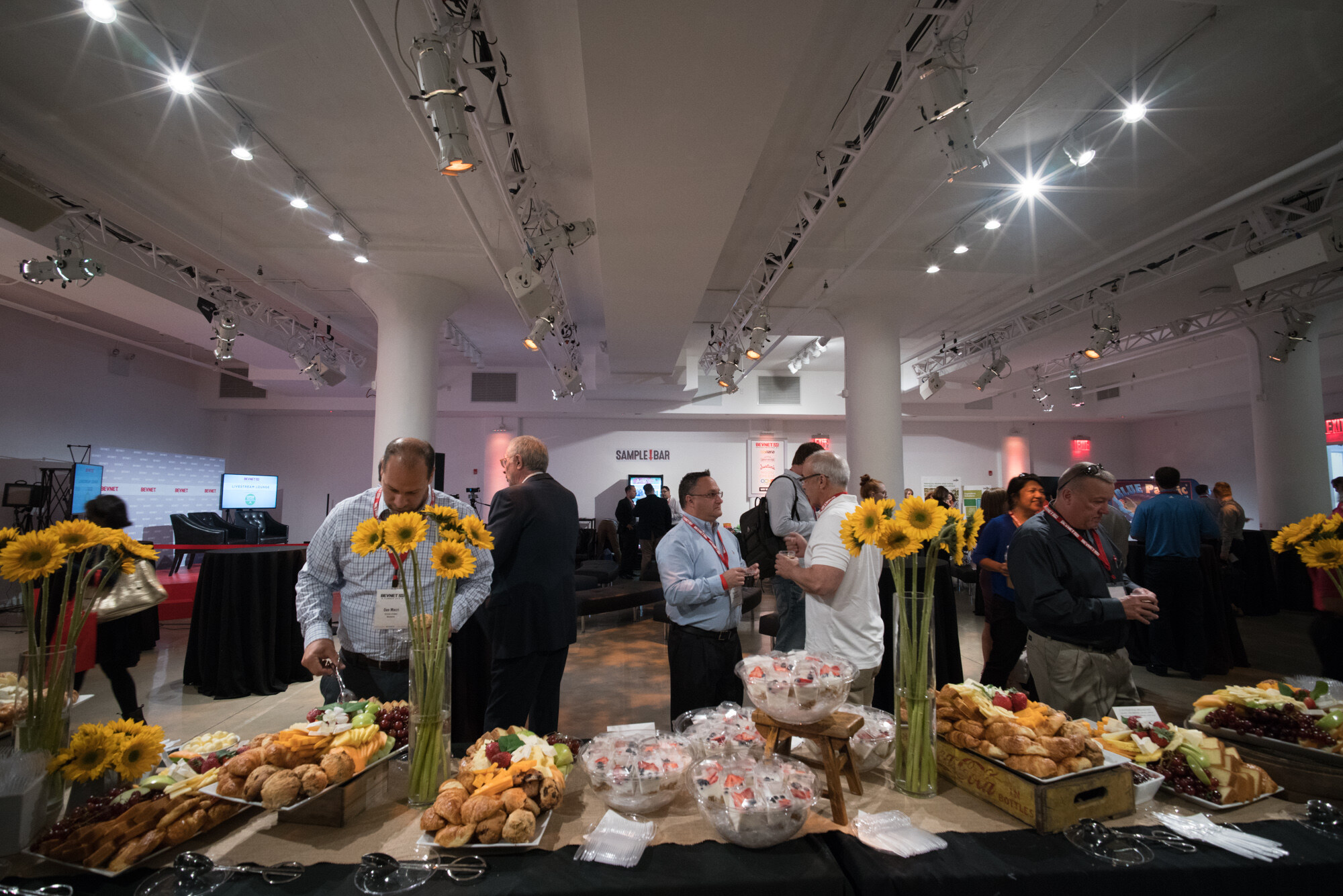 Wide-angle view of a buffet of pastries and fruit with sunflowers in the foreground, attendees talking, and a Sample Bar sign on the far wall.