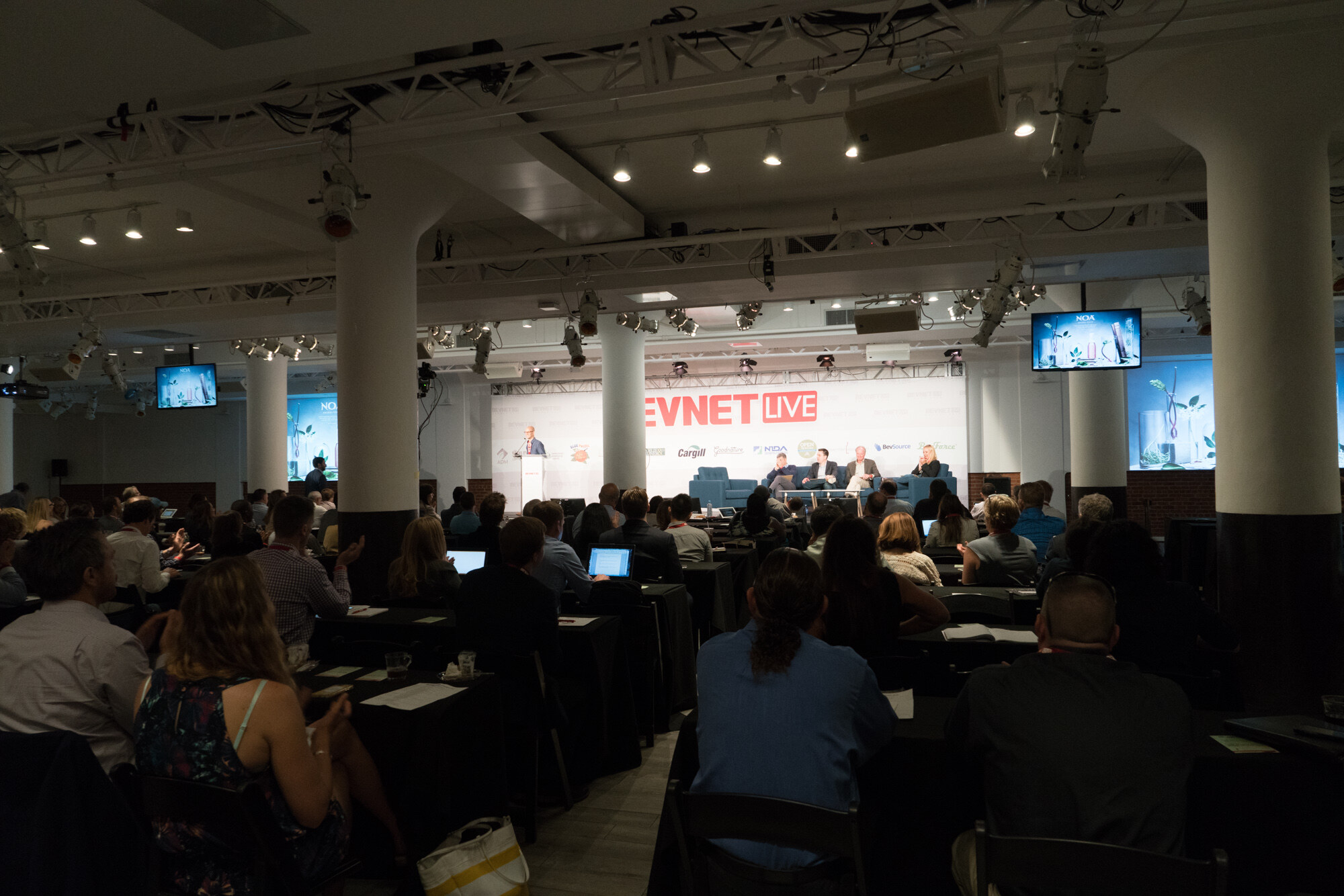 Dark room with white columns and stage lights, rows of seated attendees with laptops, and a panel on stage under a BevNET Live sign.