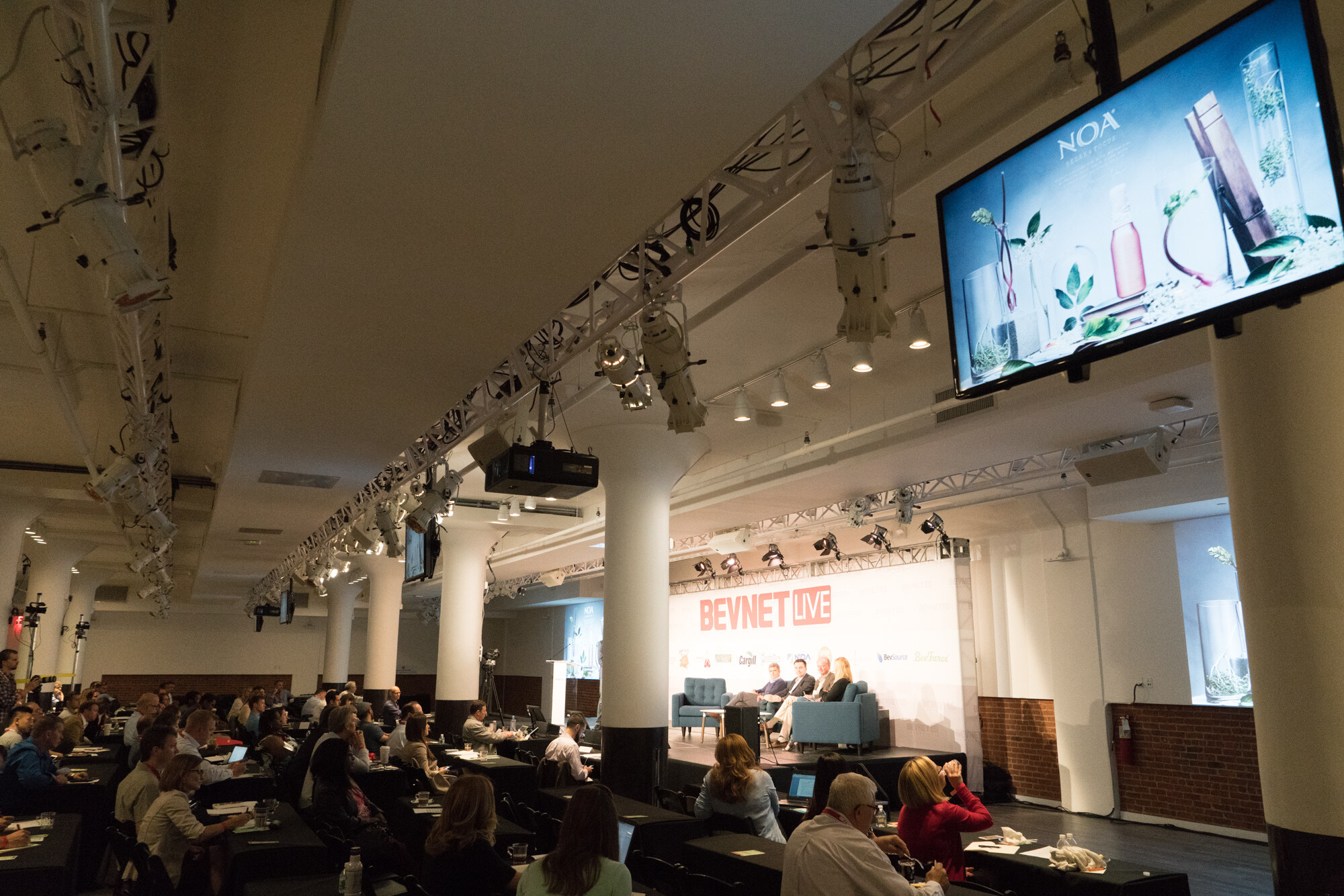 Wide-angle view of the main hall with rows of tables, a panel on stage under the BevNET Live sign, and a monitor reading NOA mounted at the right.