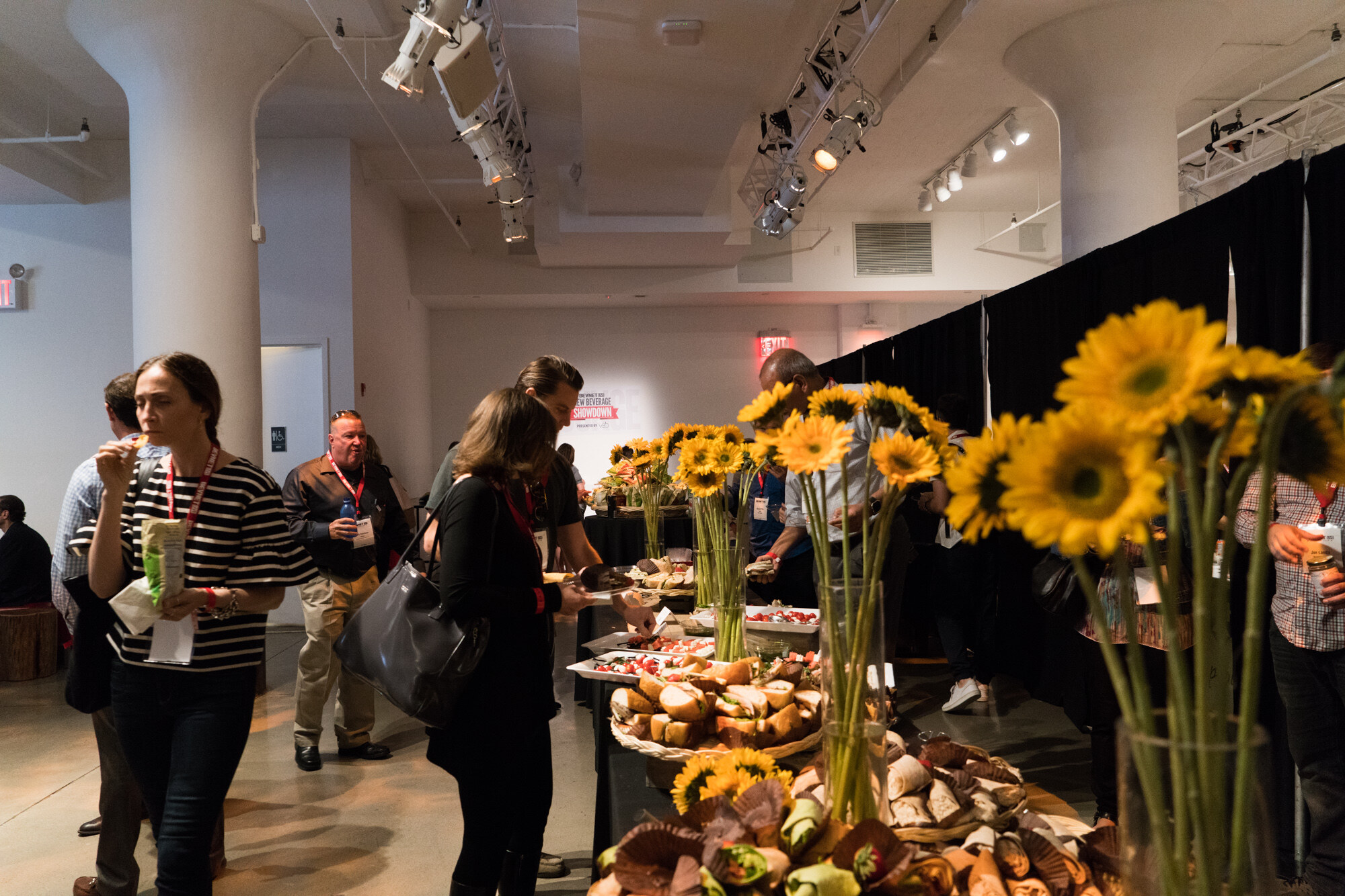 Attendees with lanyards at a long black-draped buffet table of sandwiches and pastries with vases of sunflowers, with a New Beverage Showdown sign on the far wall.