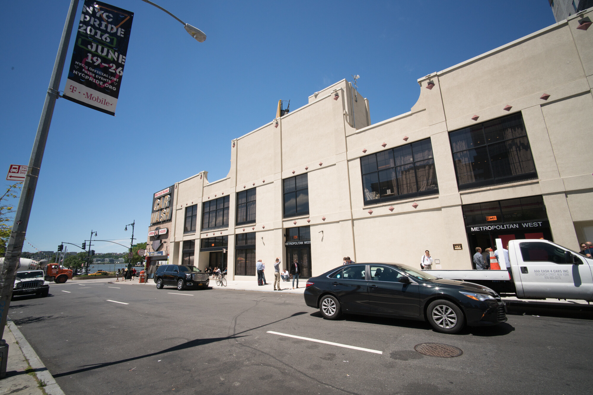 Street view of a low cream building with large dark windows and a Metropolitan West sign, a black car and a white pickup parked in front, and an NYC Pride 2016 banner at left.