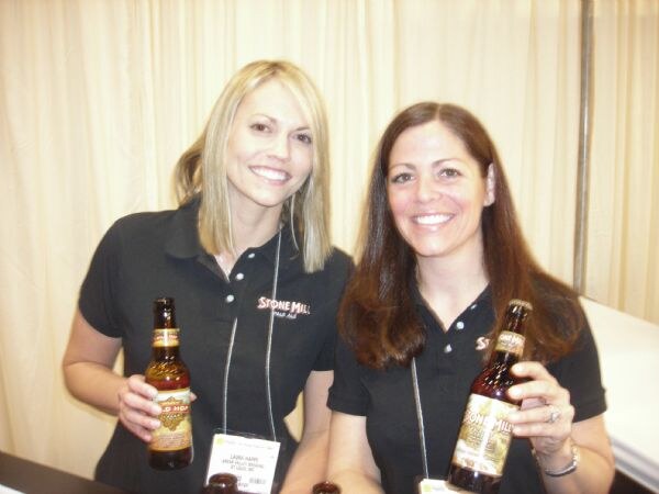 Two smiling women in black Stone Mill polo shirts each hold a bottle of Stone Mill beer in front of a cream-colored curtain.