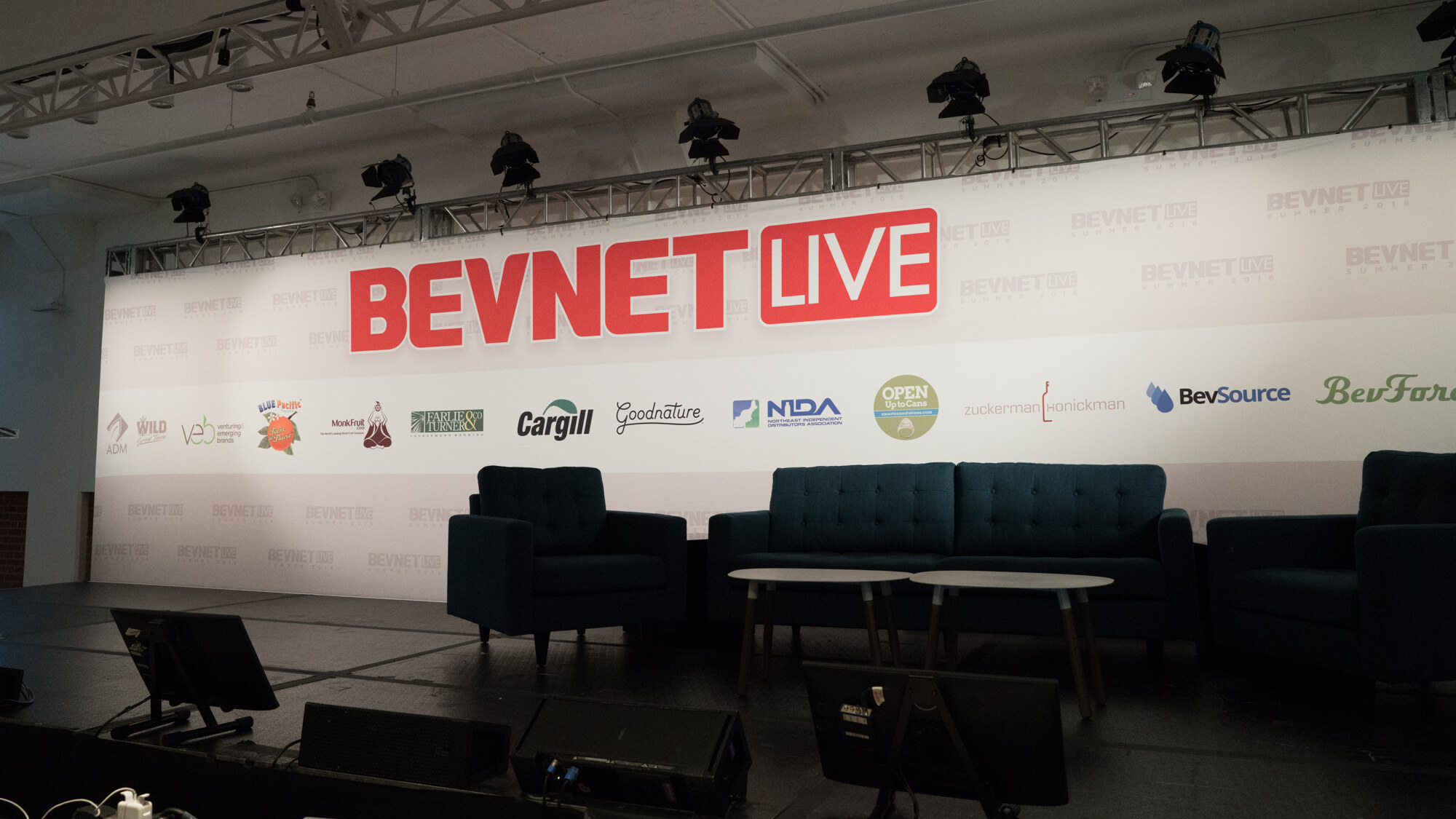 Empty stage with a dark teal sofa and two armchairs in front of a white backdrop with a red BevNET Live logo and sponsor logos including Cargill, Goodnature, NIDA, BevSource and BevForce.