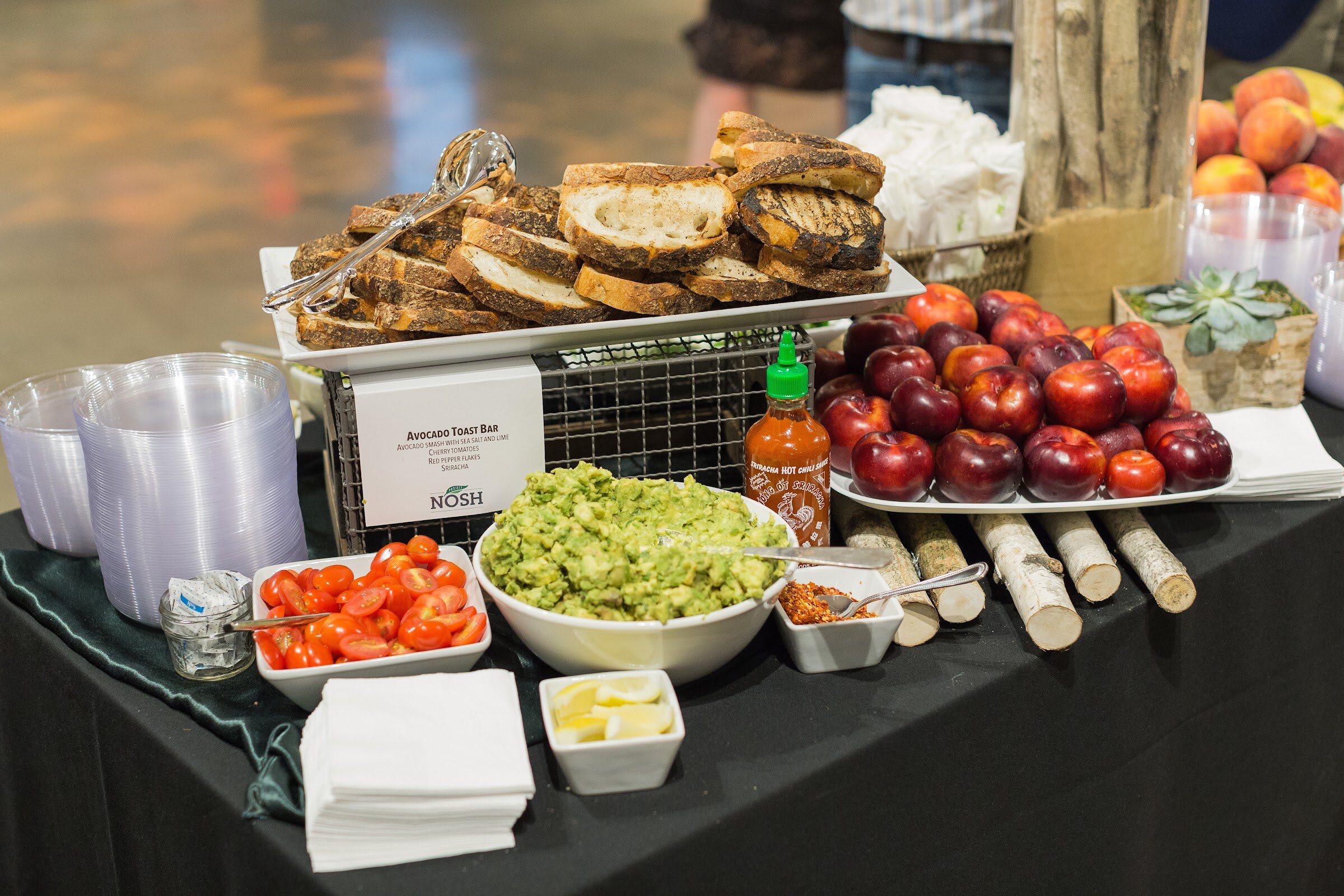 Black-draped buffet table with a platter of grilled bread, a bowl of mashed avocado, cherry tomatoes, lemon wedges, a bottle of sriracha and plums, with a card reading Avocado Toast Bar and a Nosh logo.