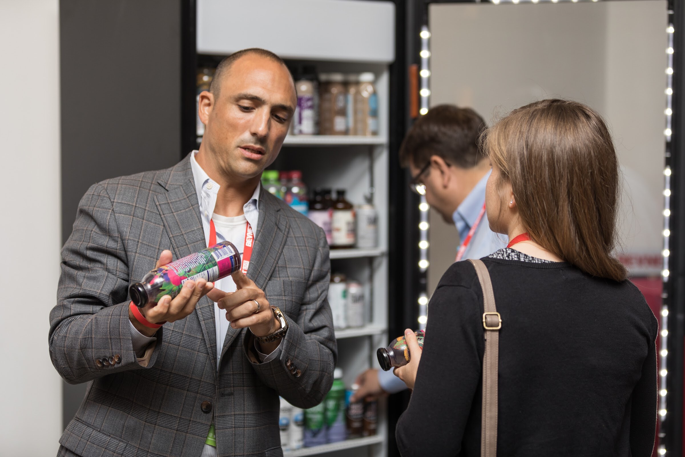 Man in a gray plaid blazer holding a bottle and pointing at its label while a woman with a shoulder bag holds another bottle, in front of a lit drink cooler.