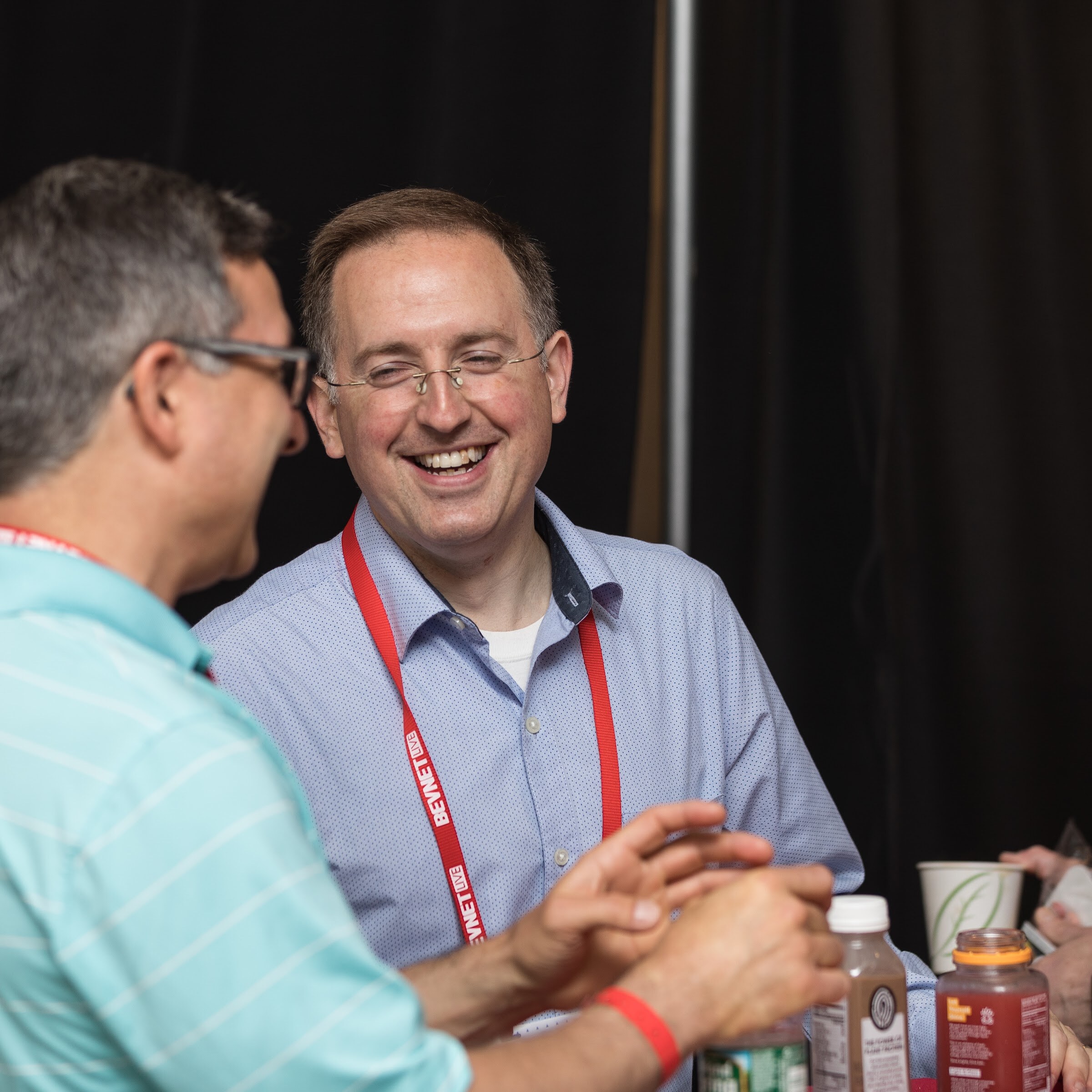 Smiling man in glasses and a blue dotted shirt with a red BevNET Live lanyard laughing while a man in a teal striped polo gestures in the foreground.