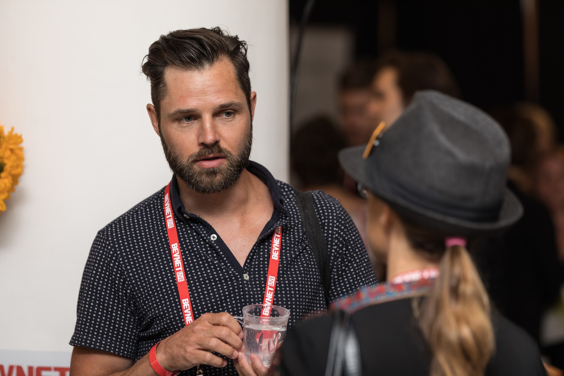 Bearded man in a navy polka-dot shirt and red BevNET Live lanyard holding a clear cup, with a woman in a gray hat in the foreground.
