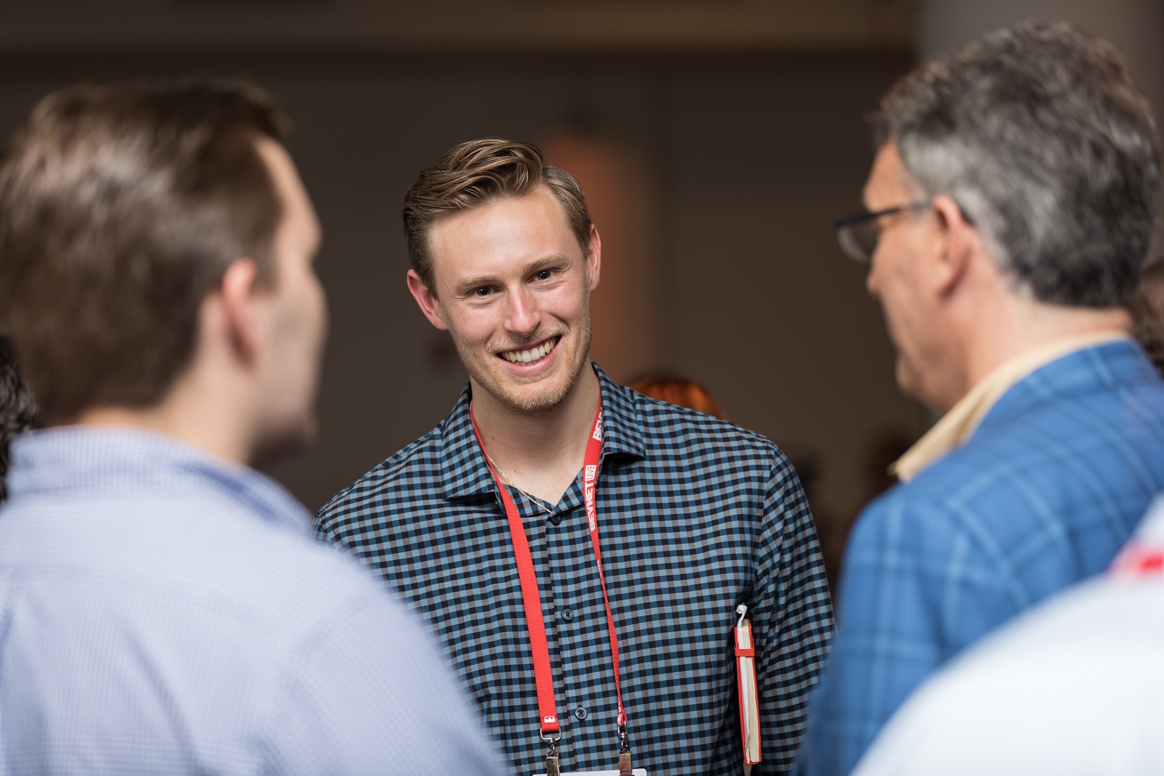 Smiling young man in a navy checked shirt and red lanyard listening to two people seen from behind.
