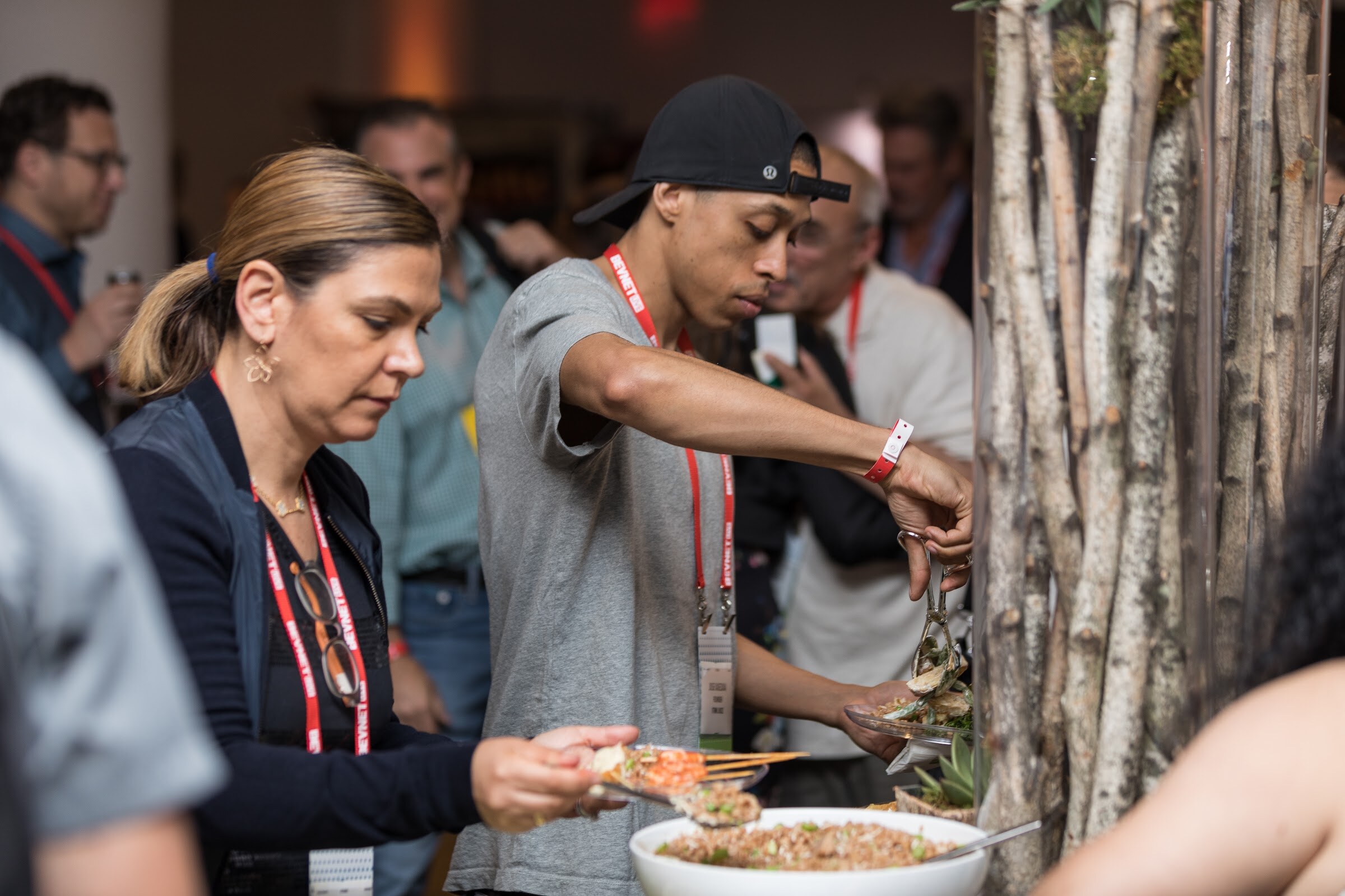 Man in a gray T-shirt and backward black cap using tongs at a buffet next to a woman in a navy vest, with a birch-branch centerpiece at right.