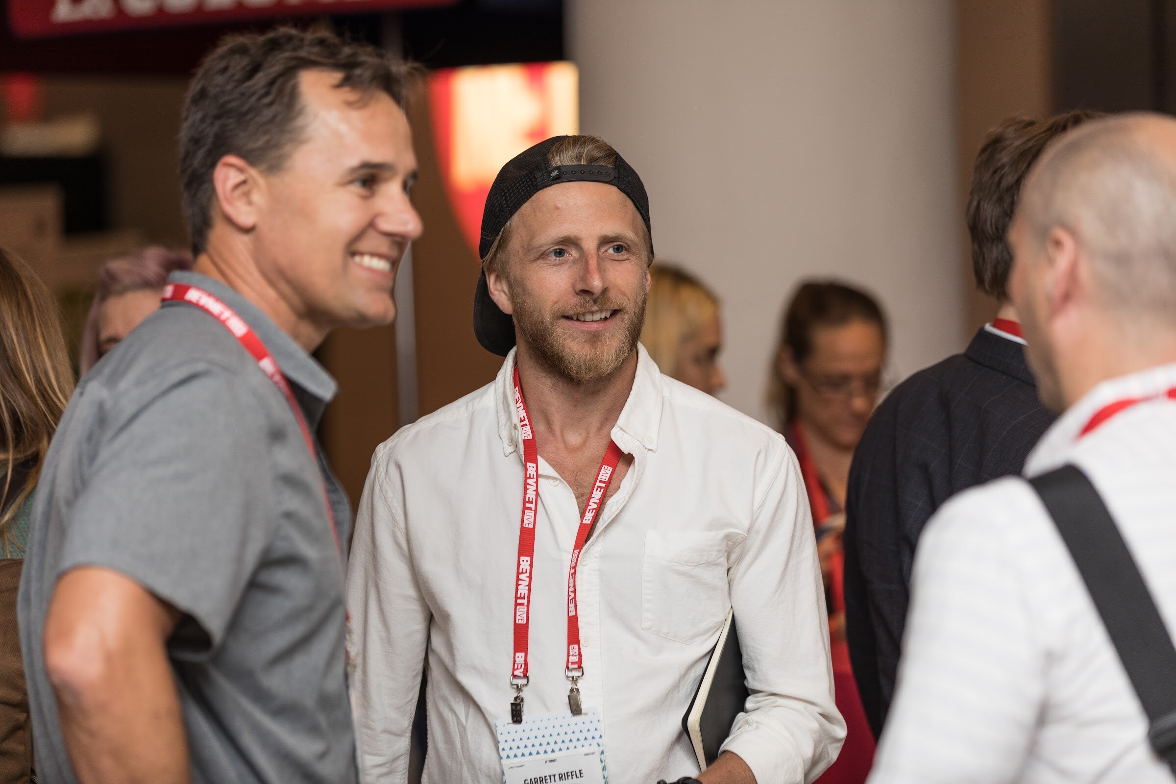 Man in a white shirt, backward black cap and red BevNET Live lanyard smiling at a man in a gray polo, with other attendees blurred behind.
