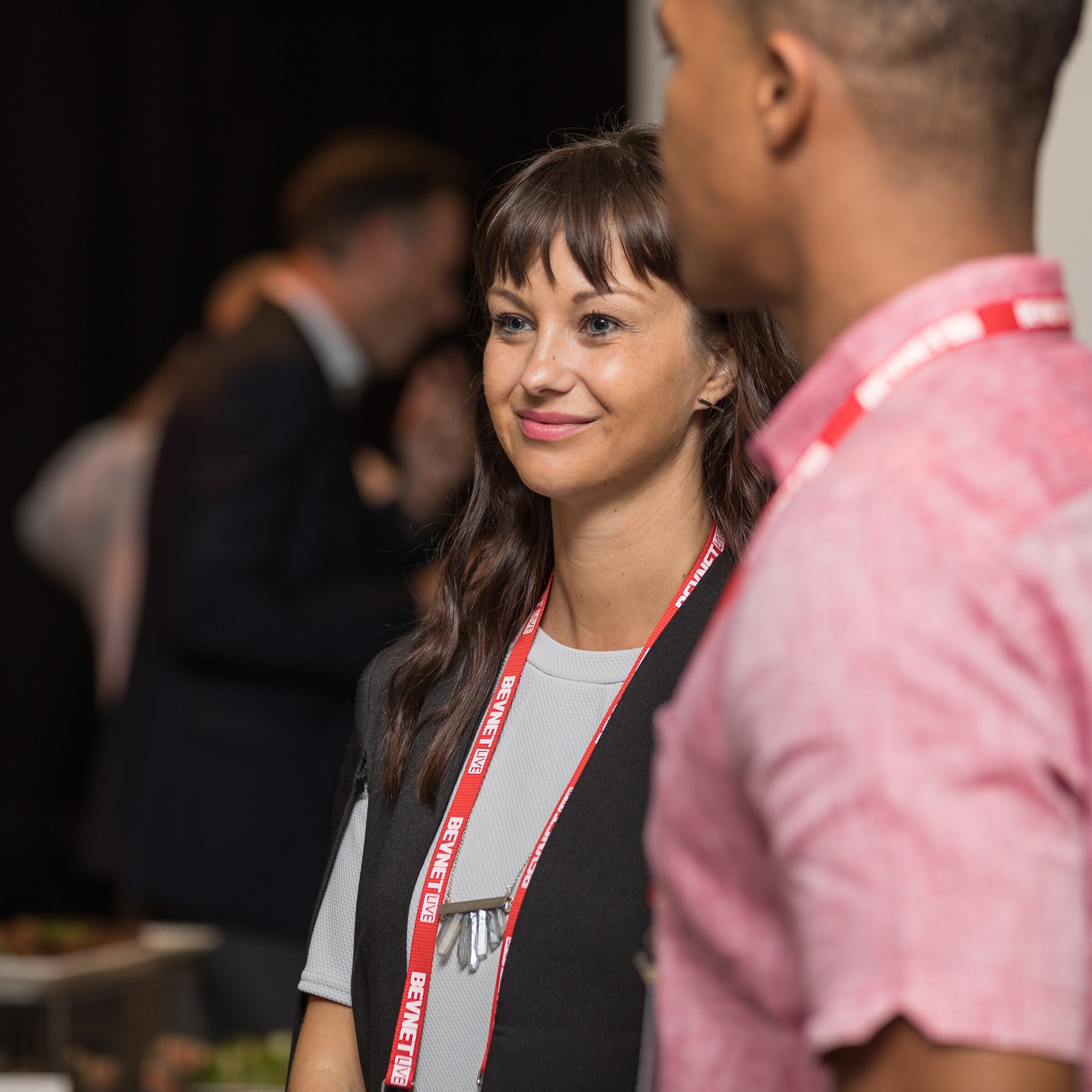 Smiling woman with dark hair and a black vest over a gray top and a red BevNET Live lanyard, with the shoulder of a man in a pink shirt in the foreground.