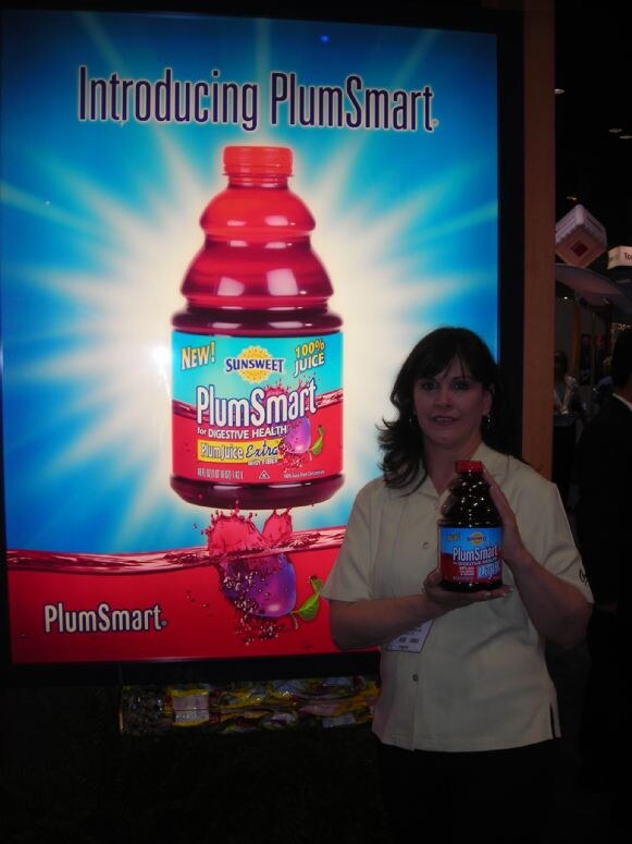 A woman with dark hair holds a bottle of Sunsweet PlumSmart juice beside a large lit sign reading "Introducing PlumSmart" that shows a bottle of the juice.