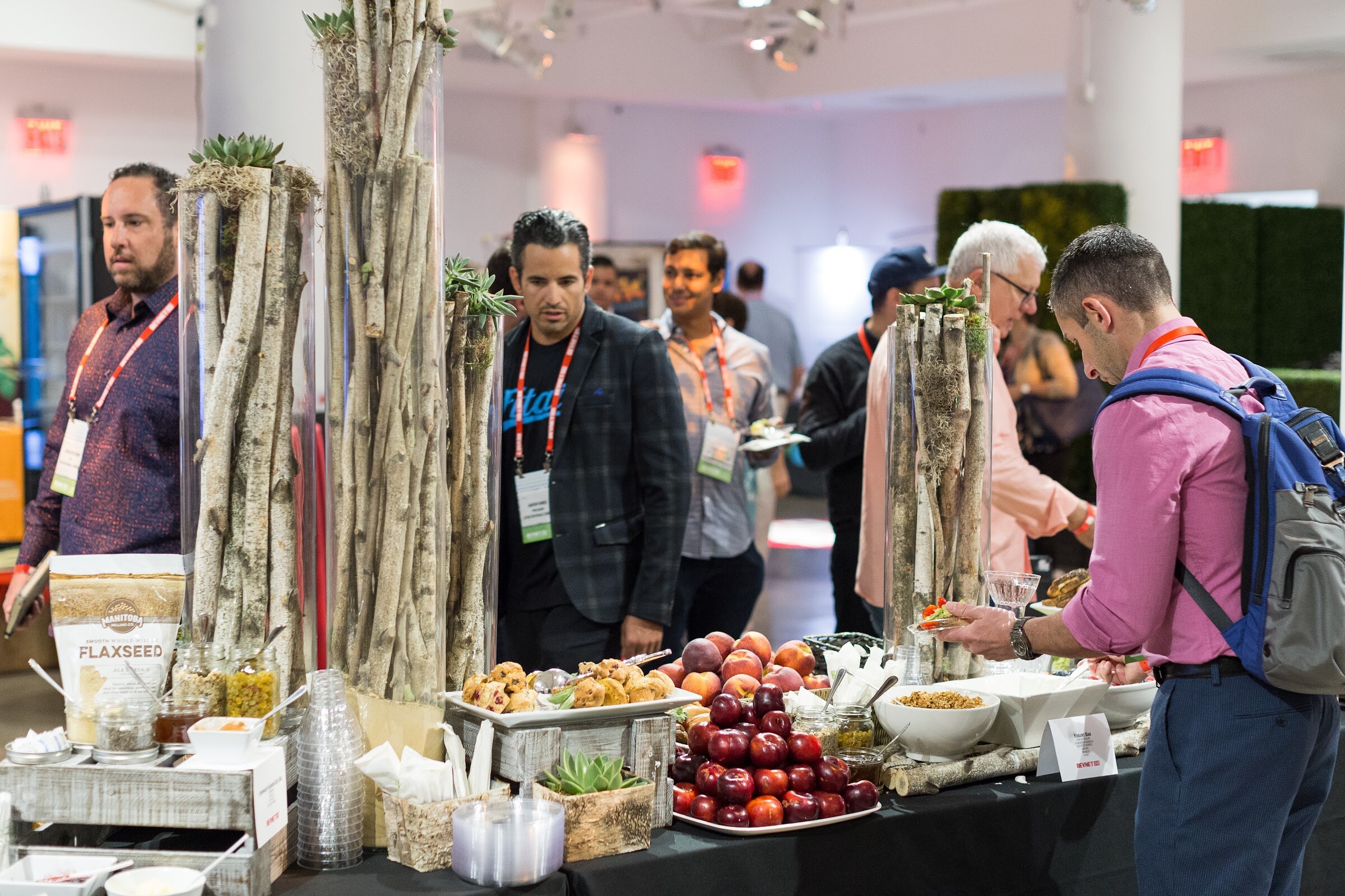 Attendees at a buffet table with plums, nectarines, flaxseed and snacks in front of tall birch-branch vases, with a man in a pink shirt and backpack at right.