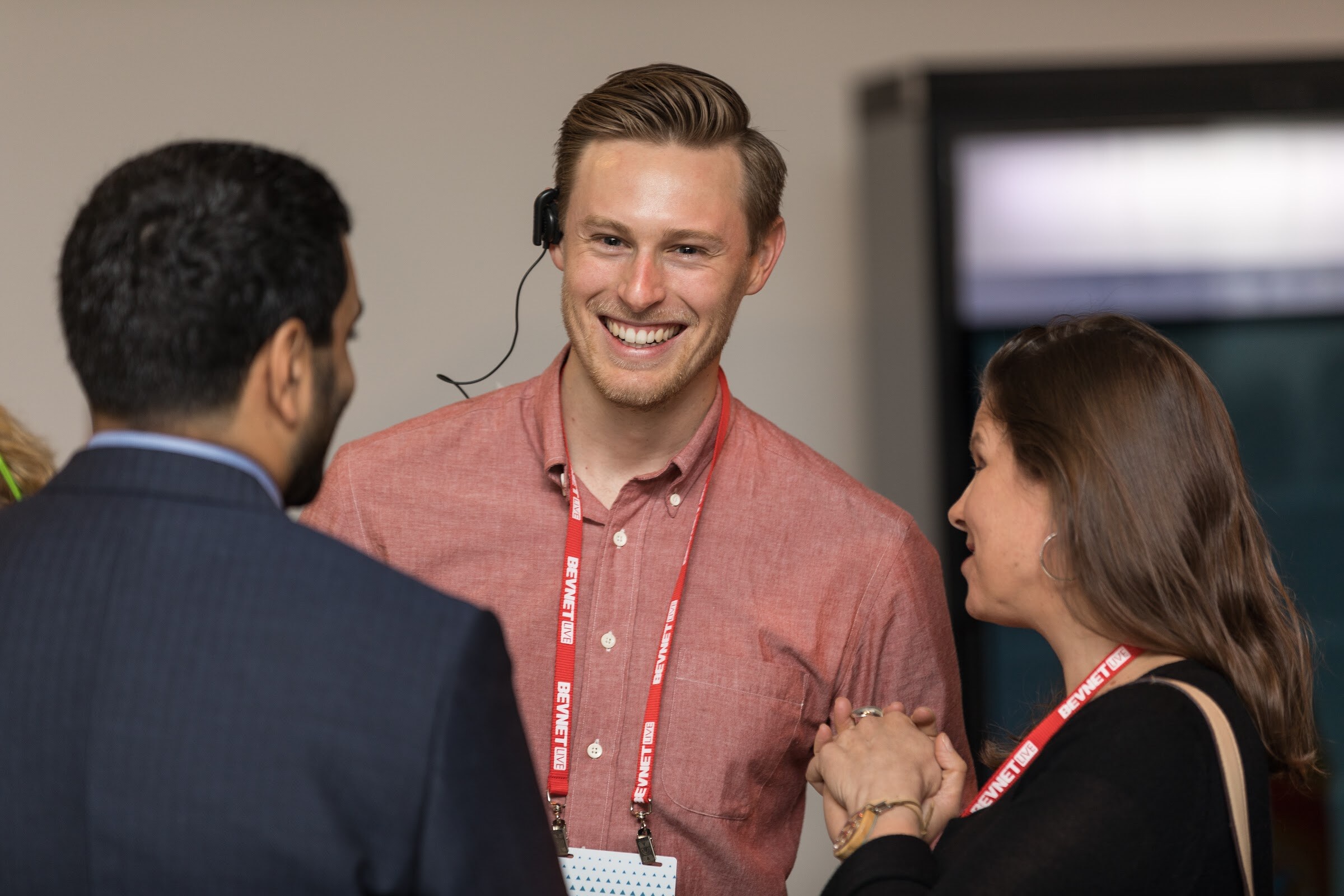 Smiling man in a coral shirt and red lanyard with a single earpiece talking with a man in a navy jacket and a woman in black.