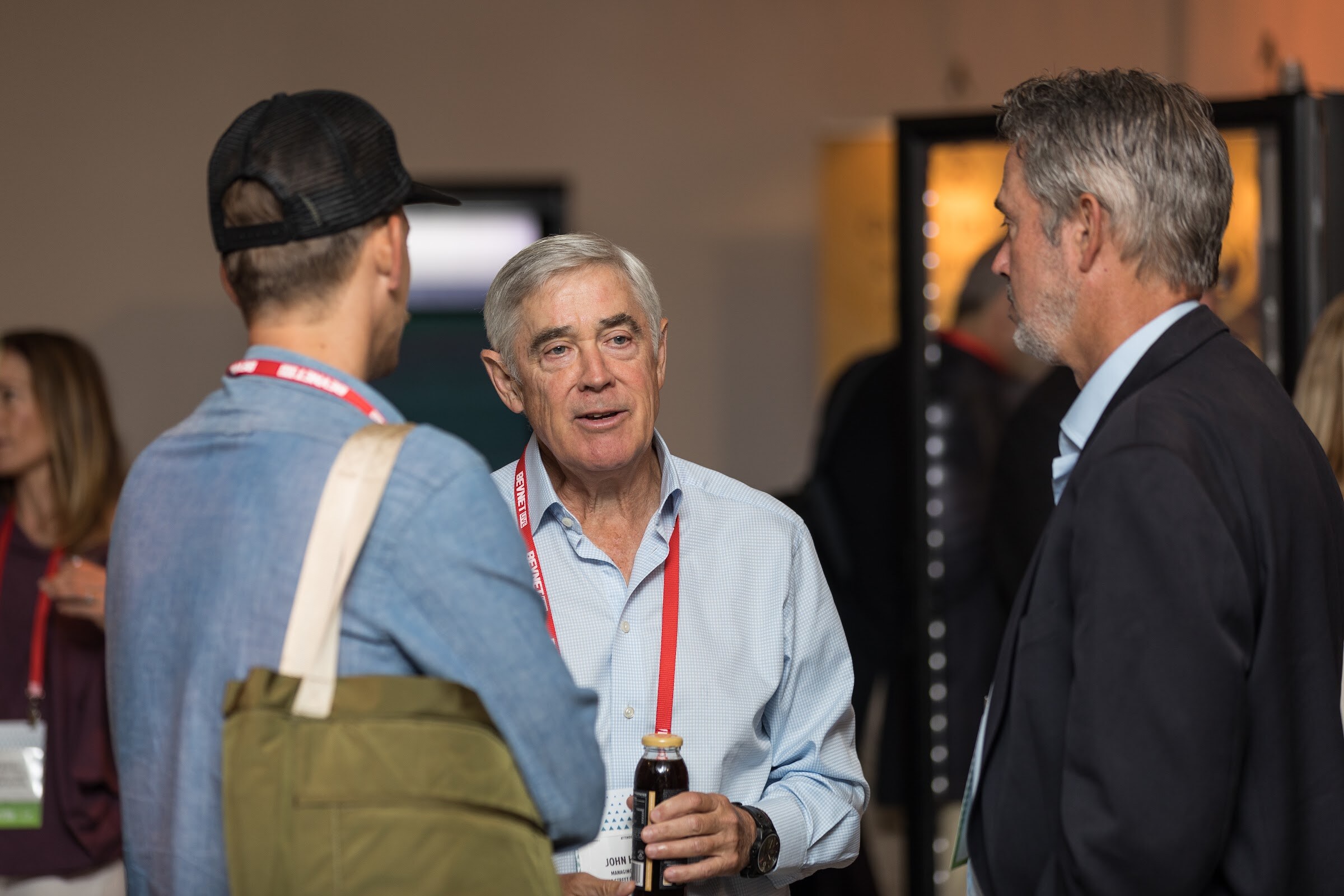 A white-haired man in a light blue shirt holding a bottle of dark juice talking with a man in a black blazer and another in a denim jacket and black cap seen from behind.