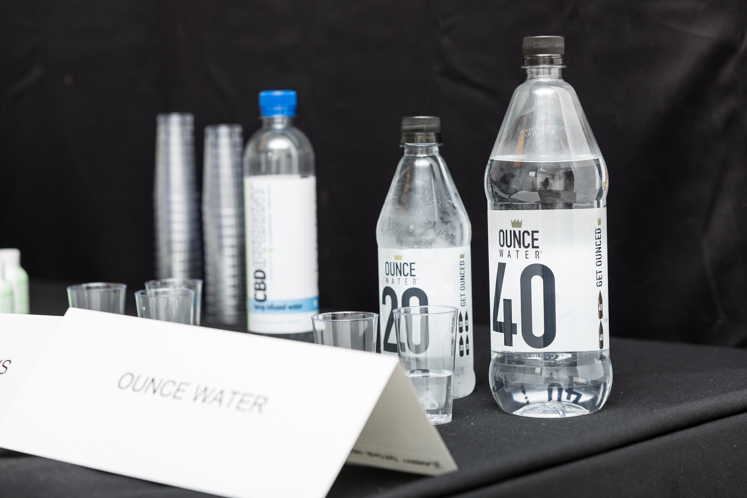 Bottles labeled OUNCE WATER 20 and 40 and a CBD-infused water bottle on a black tablecloth with small plastic tasting cups and a card reading OUNCE WATER.