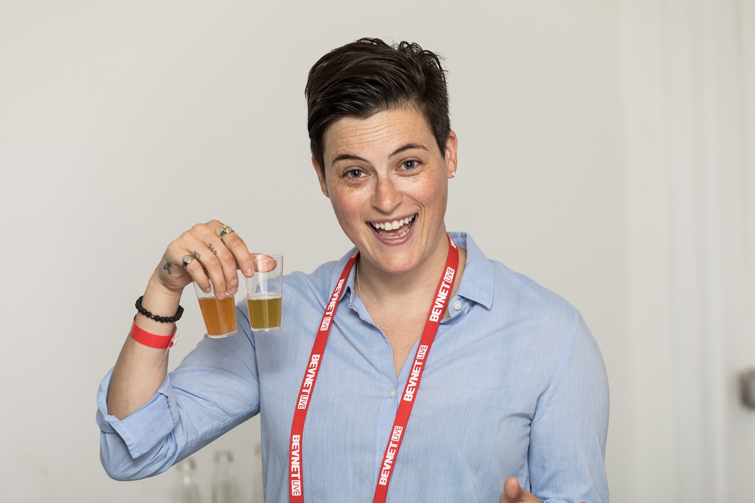 Person with short dark hair in a light blue shirt and red BEVNET LIVE lanyard, smiling and holding two small cups of amber drinks.