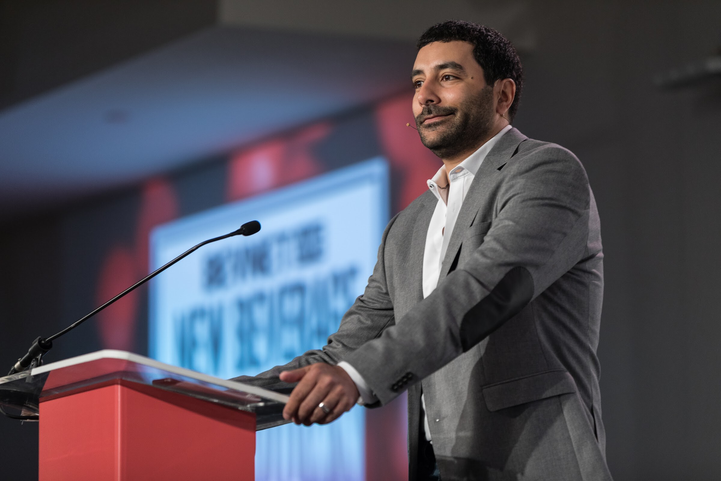 Smiling man in a gray blazer with a headset microphone at a red podium, with a blurred slide reading New Beverage Showdown behind him.