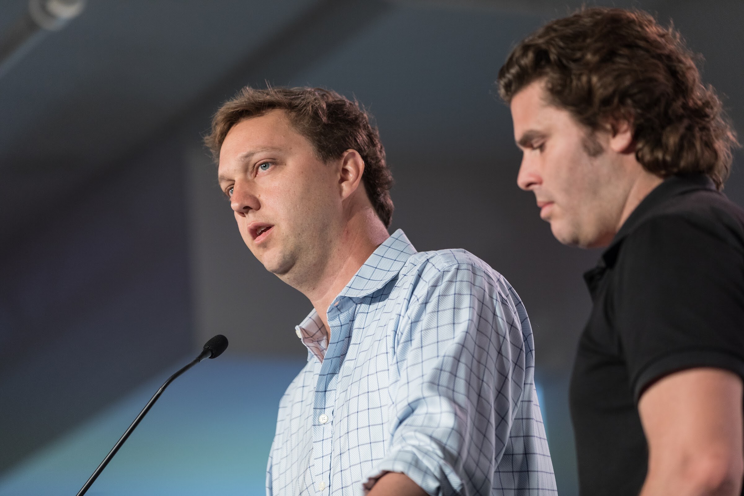 Close view of a man in a checked shirt speaking at a microphone while a man in a black shirt looks down beside him.