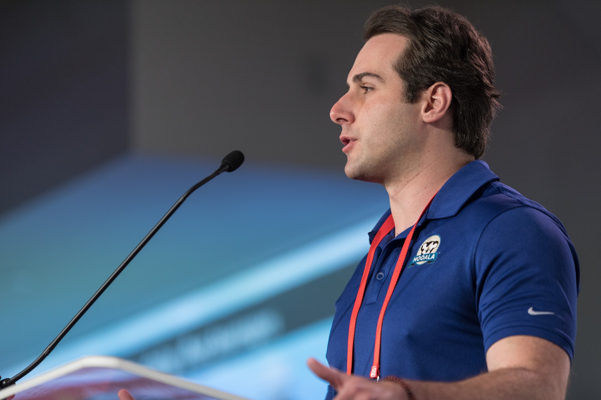 Man in a blue polo with a Mooala logo and red lanyard speaking in profile at a podium microphone.