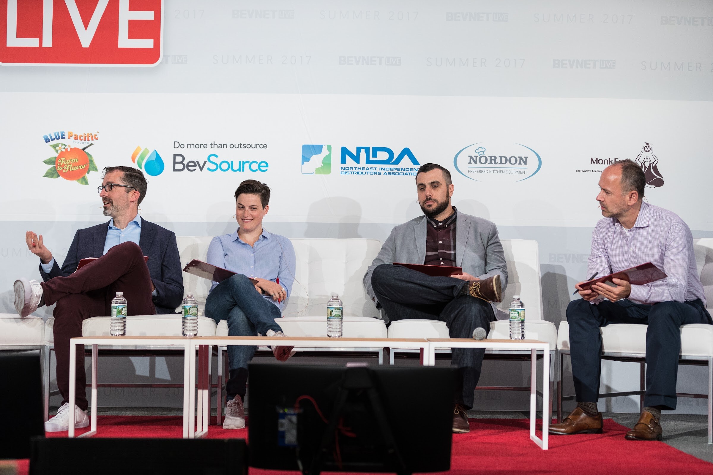 Four people seated on white lounge chairs holding red clipboards, with water bottles on low tables and BevSource, NIDA, Nordon and Monk Fruit logos behind them.