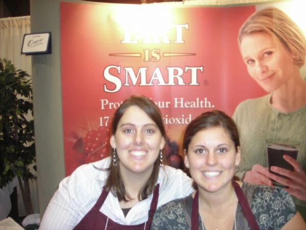 Two smiling women in maroon aprons stand in front of a red Tart is Smart banner showing berries and a woman holding a glass of dark juice.