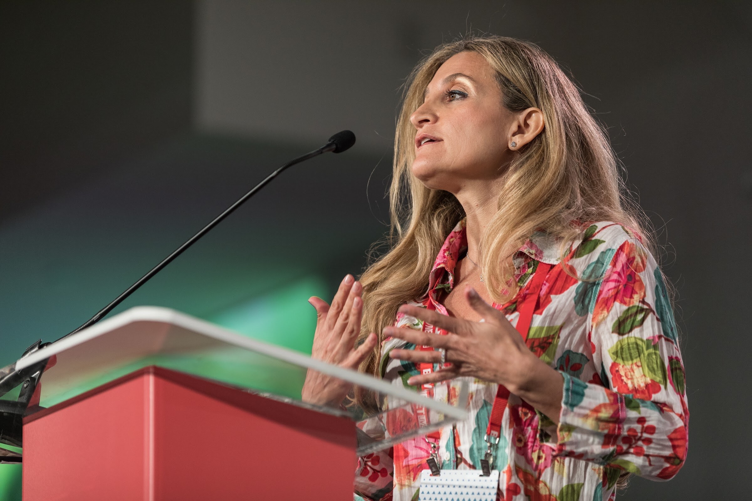 Woman with long blonde hair in a bright floral blouse and red lanyard speaking with both hands raised at a red podium.