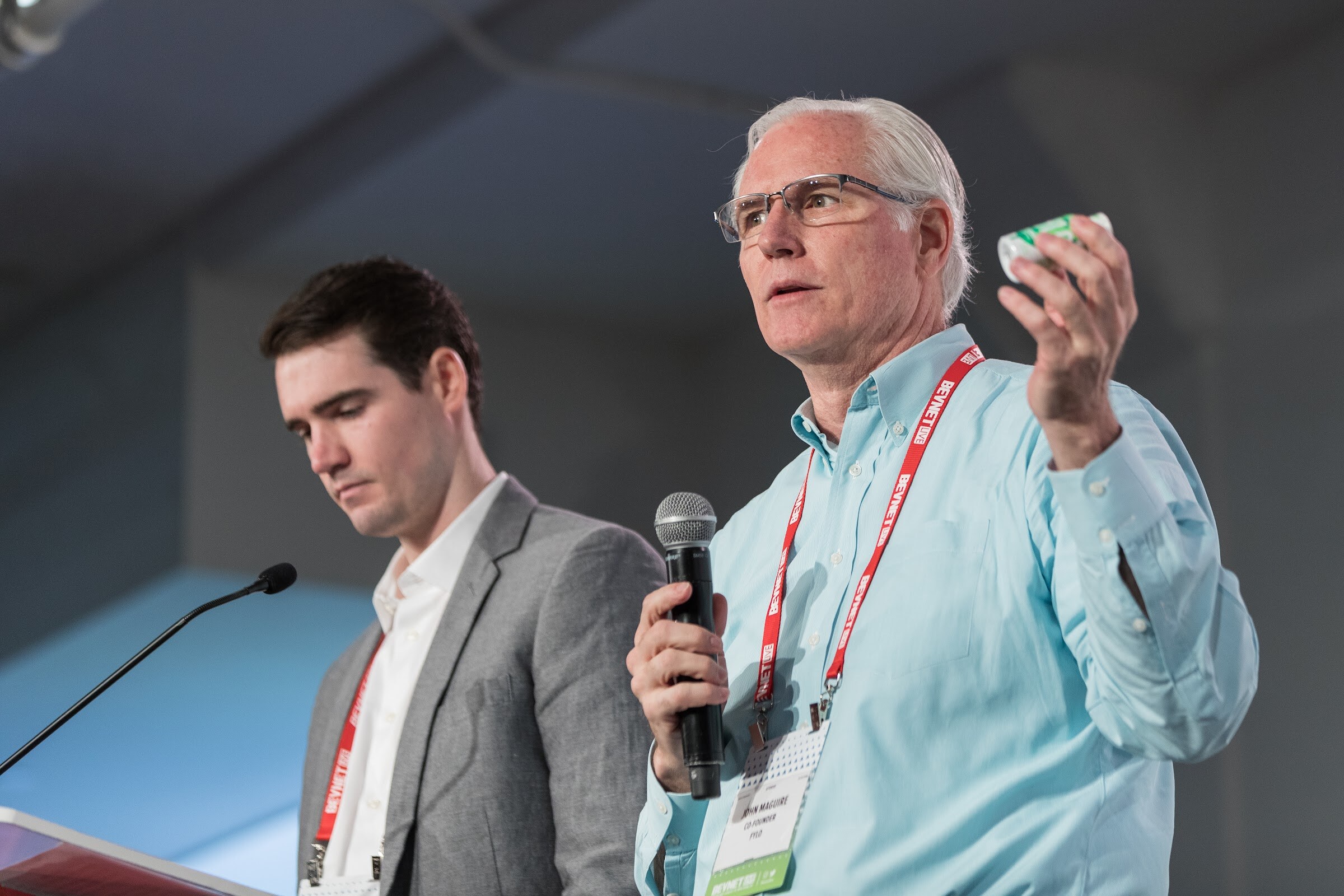 Man with white hair and glasses in a light blue shirt and red lanyard holding a microphone and a small green package, with a younger man in a gray blazer looking down beside him.