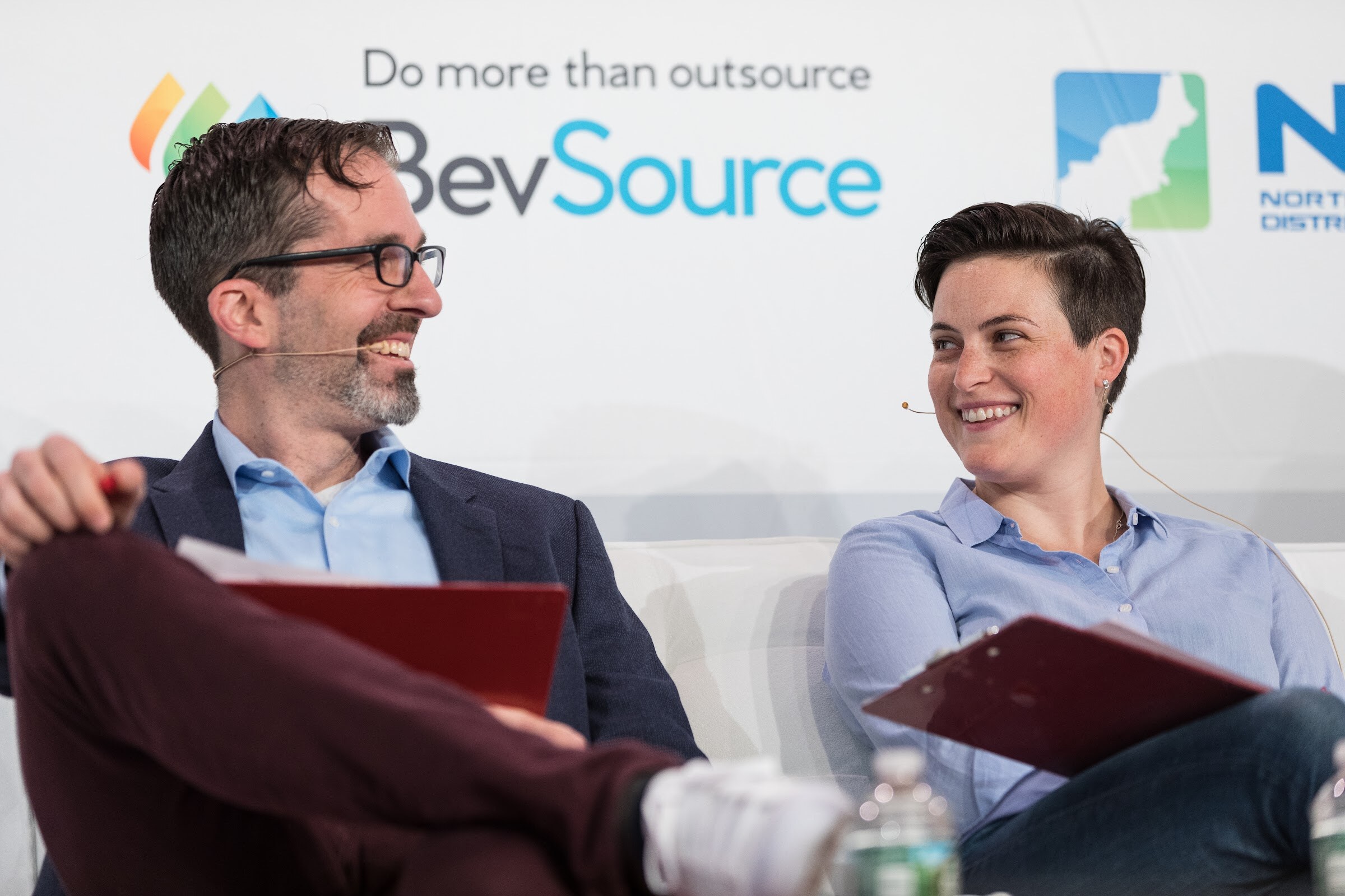 Bearded man in glasses and a blue blazer laughing with a person with short dark hair in a blue shirt, both holding red clipboards, in front of a BevSource logo.