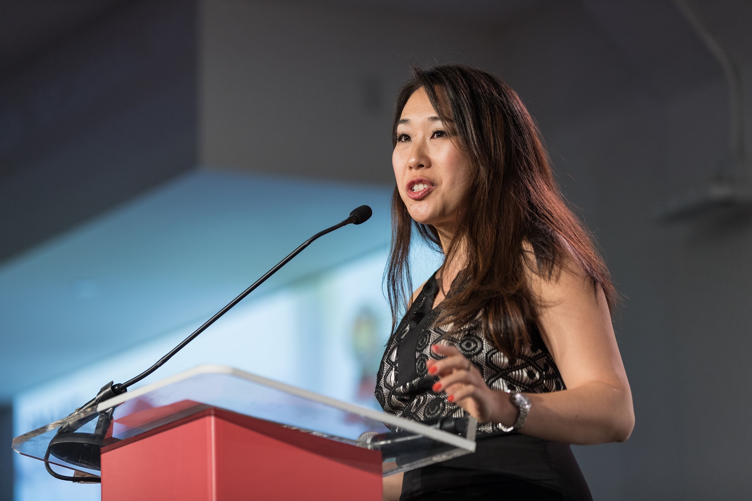 Woman with long dark hair and red nails in a black lace-trim top speaking at a clear and red podium.