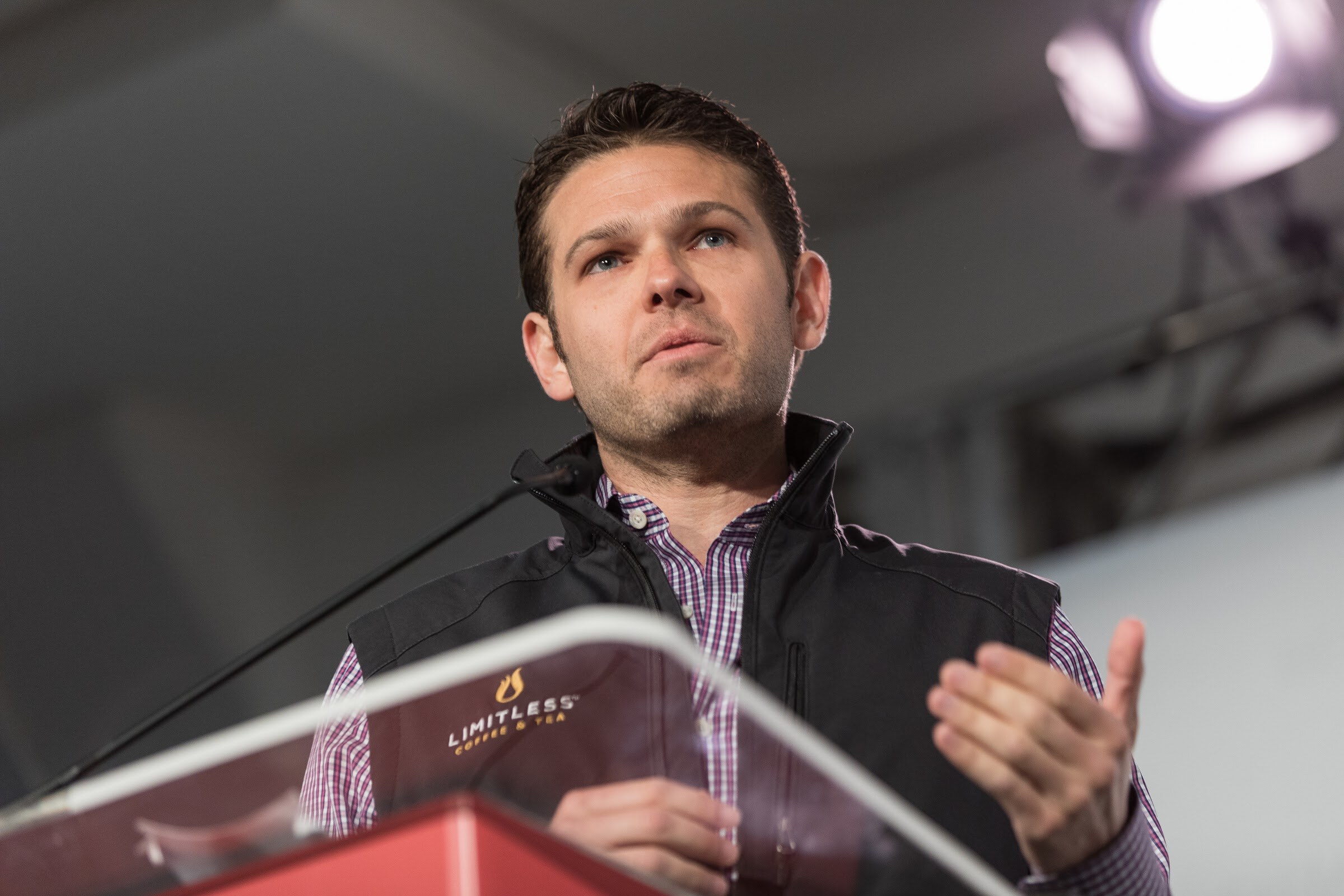 Man in a black vest over a plaid shirt speaking at a clear podium carrying a Limitless Coffee and Tea logo.