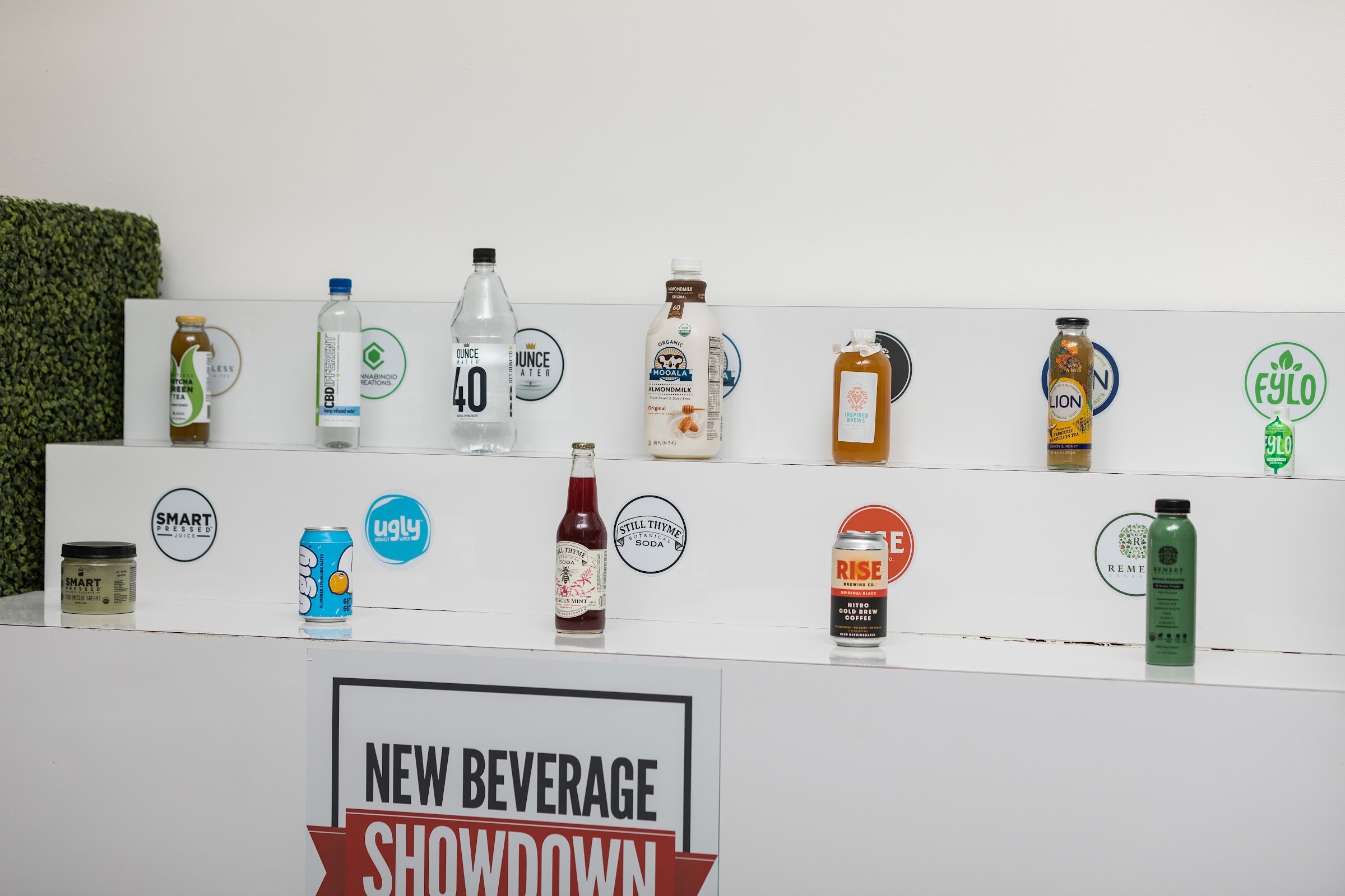 Tiered white shelves holding bottles and cans including Ounce Water, Mooala, Ugly, Still Thyme Soda, Rise and Remedy, with a New Beverage Showdown sign below.