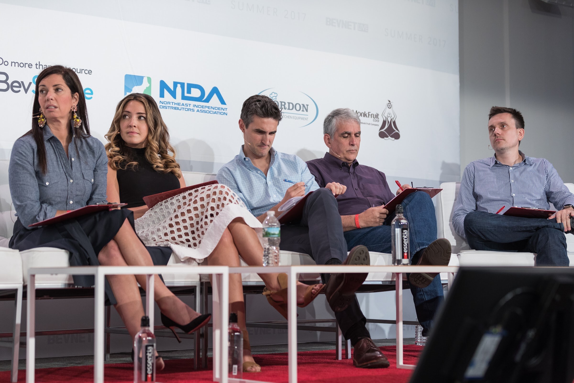 Five people seated in a row on white lounge chairs holding red clipboards, with Essentia water bottles on low tables and NIDA, Nordon and Monk Fruit logos behind them.