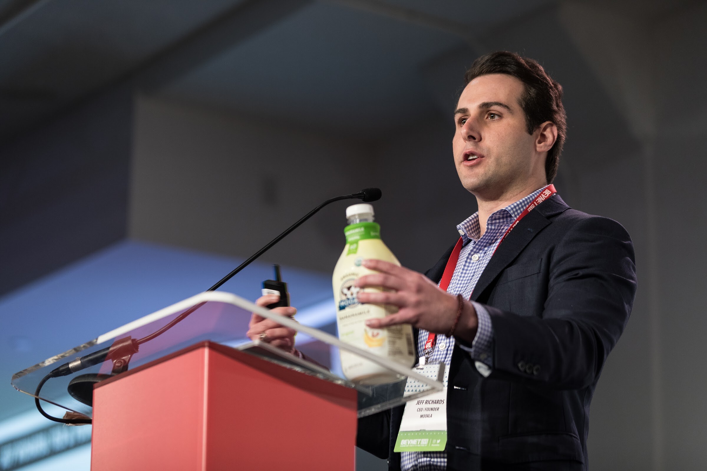 Man in a navy blazer and checked shirt with a name badge holding a bottle of Mooala plant-based milk beside a red podium and microphone.