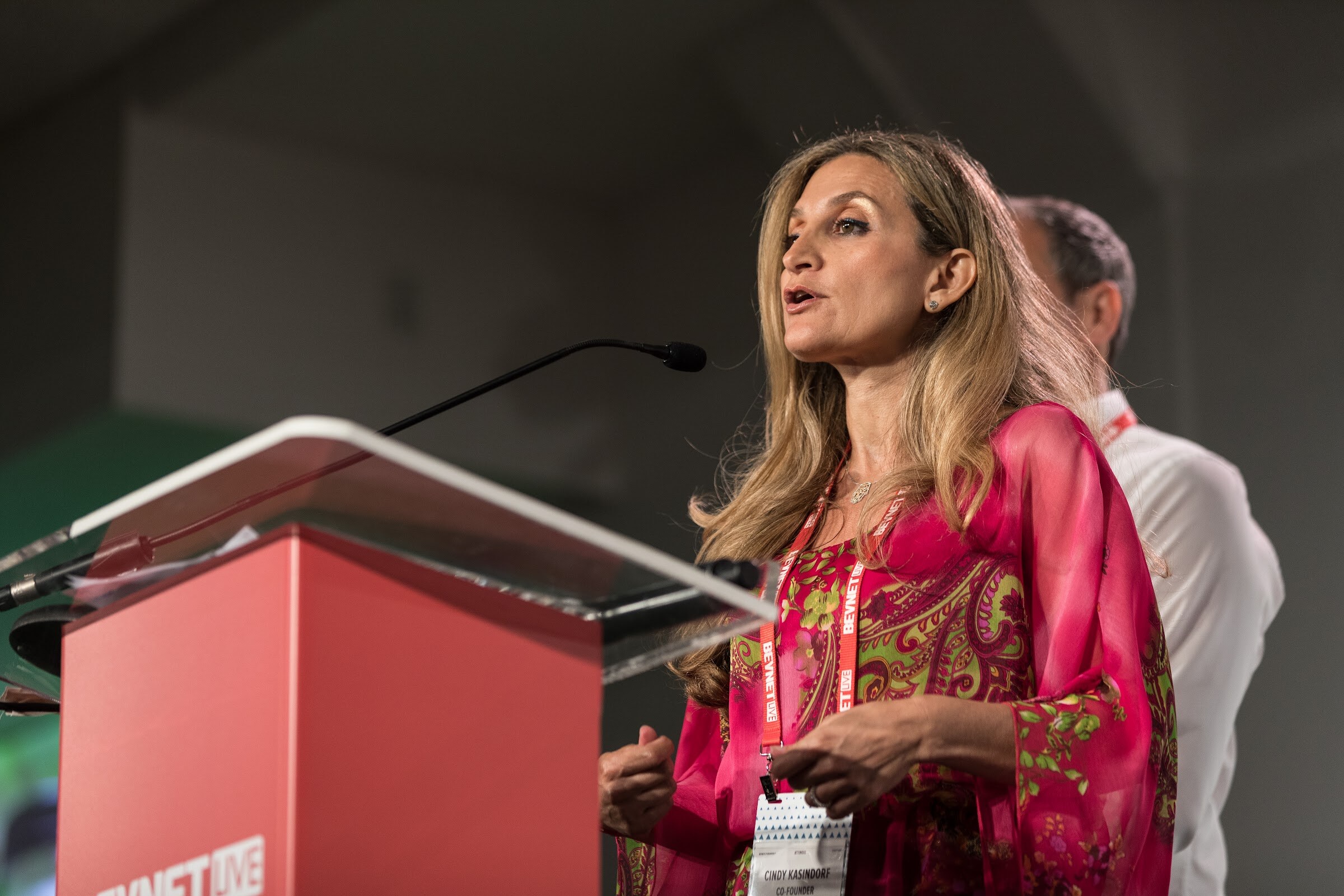 Woman with long blonde hair in a pink paisley top and a Remedy Organics badge speaking at a red podium, with a man in a white shirt partly visible behind her.