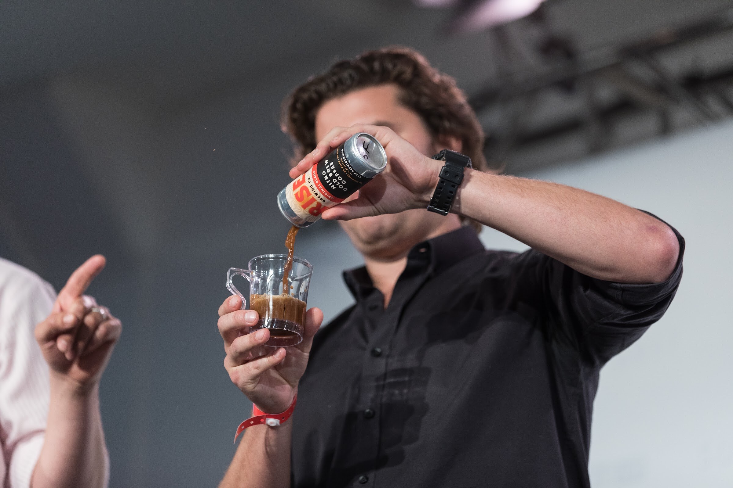 Man in a black shirt pouring coffee from a can labeled RISE into a clear sample cup, with his face hidden behind the can.