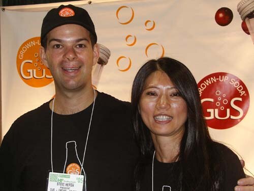 Steve Hersh, president of GuS, in a black cap and shirt, stands smiling beside a woman with long dark hair in front of a GuS Grown-Up Soda banner.