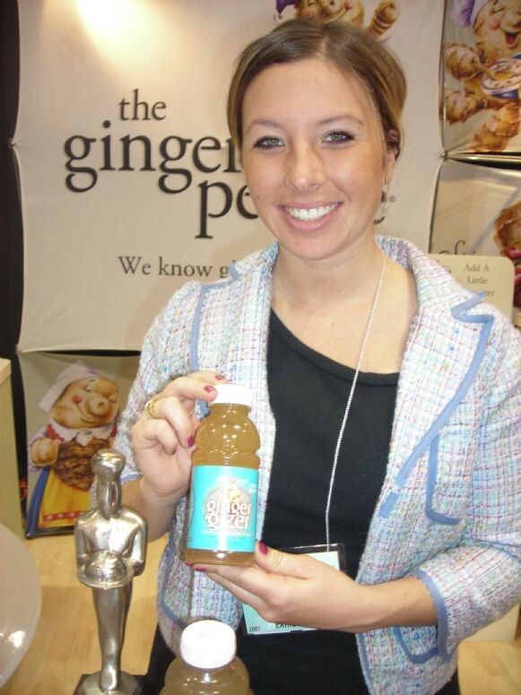 A smiling woman holds a bottle of Ginger Gizzer in front of a the ginger people banner. A small silver statuette and another bottle sit on the table in front of her.