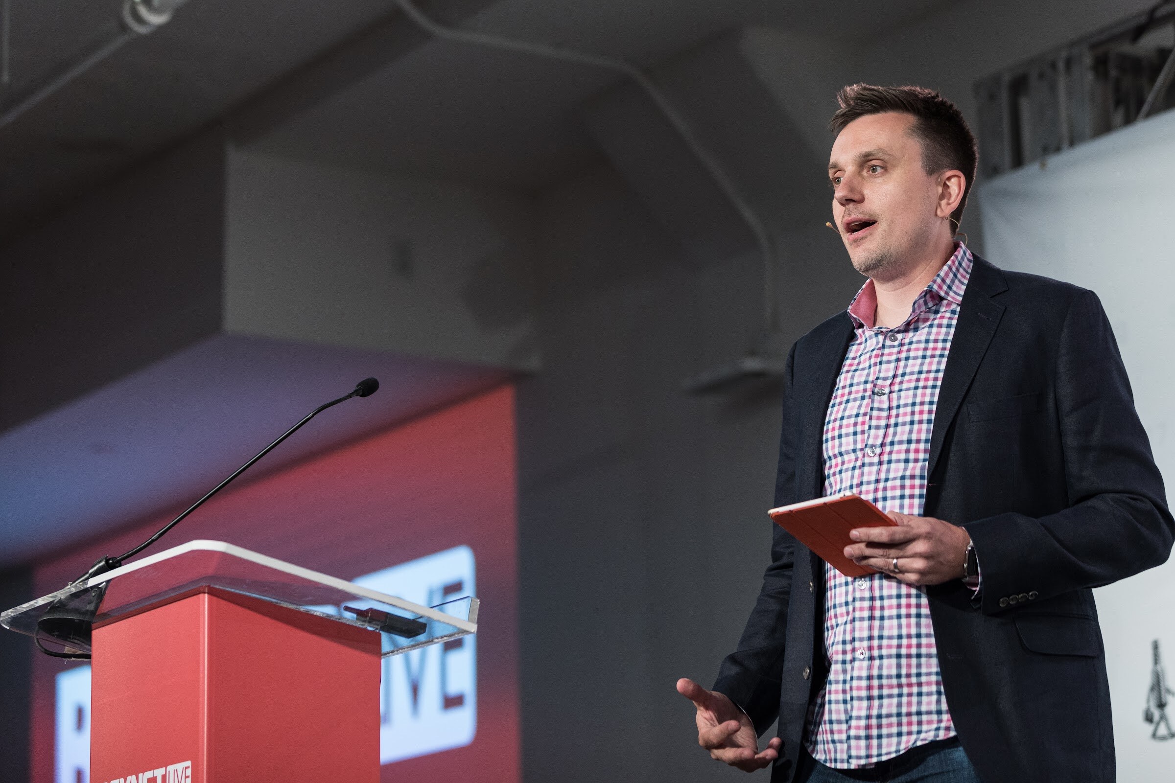 Man in a pink and navy plaid shirt and dark blazer holding an orange tablet and speaking beside a red BevNET Live podium, with a headset microphone.