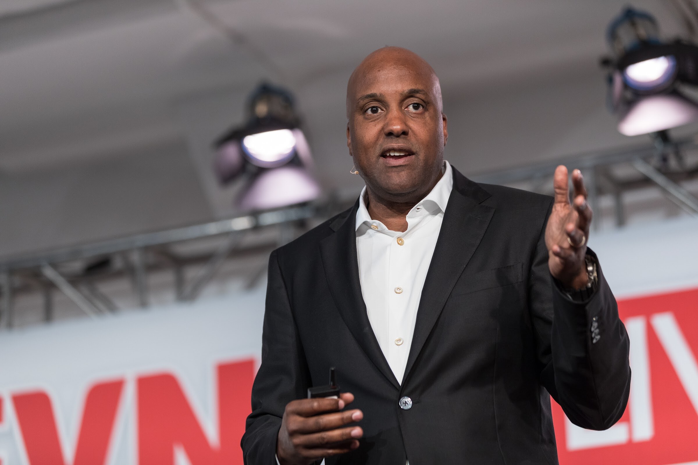 Man in a black blazer and open-collar white shirt gesturing with his right hand and holding a clicker, with stage lights and a red BEVNET LIVE sign behind him.