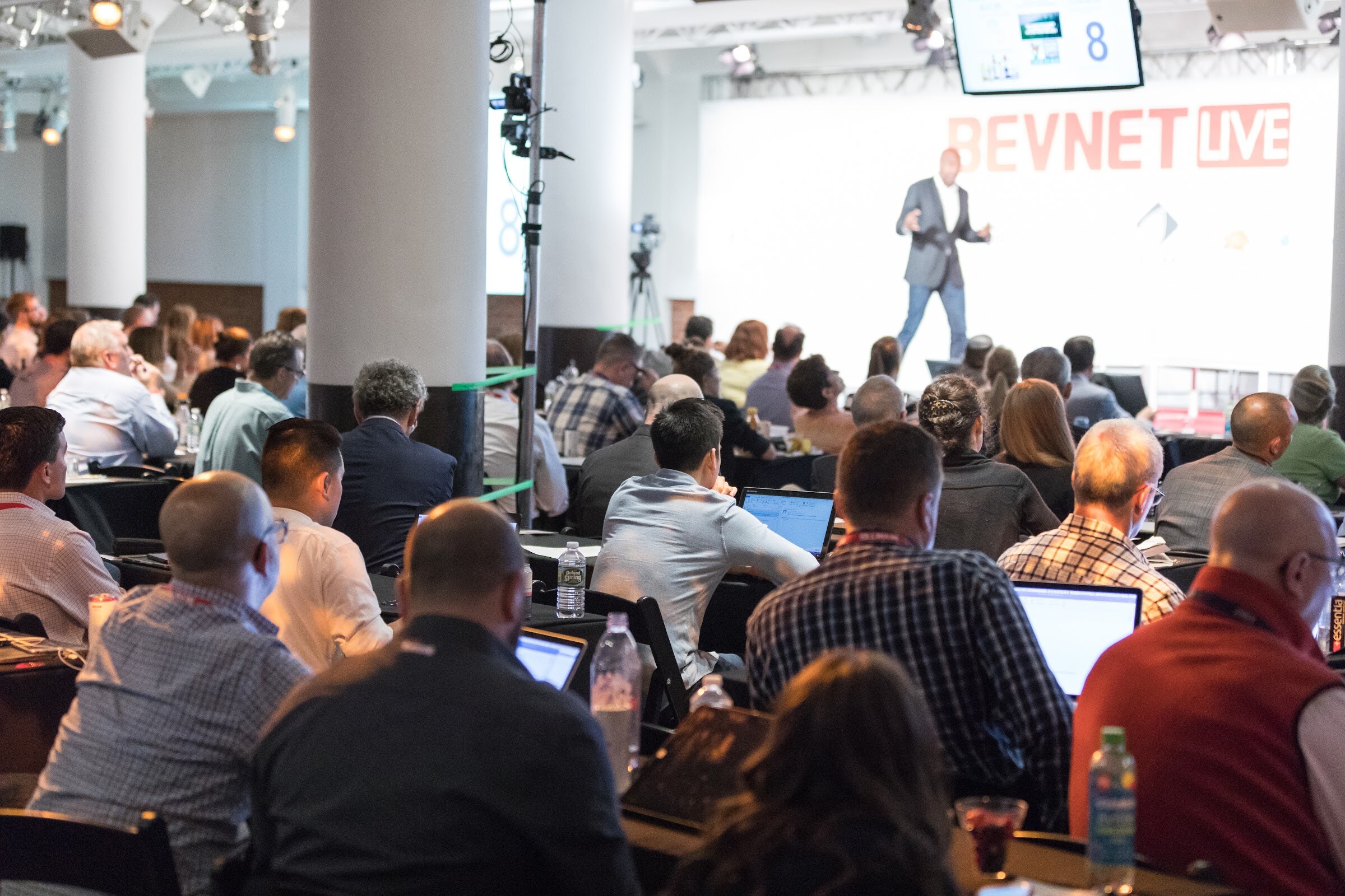 View over the shoulders of seated attendees toward a speaker on a stage in front of a BEVNET LIVE screen, with columns and a camera on a tripod in the room.