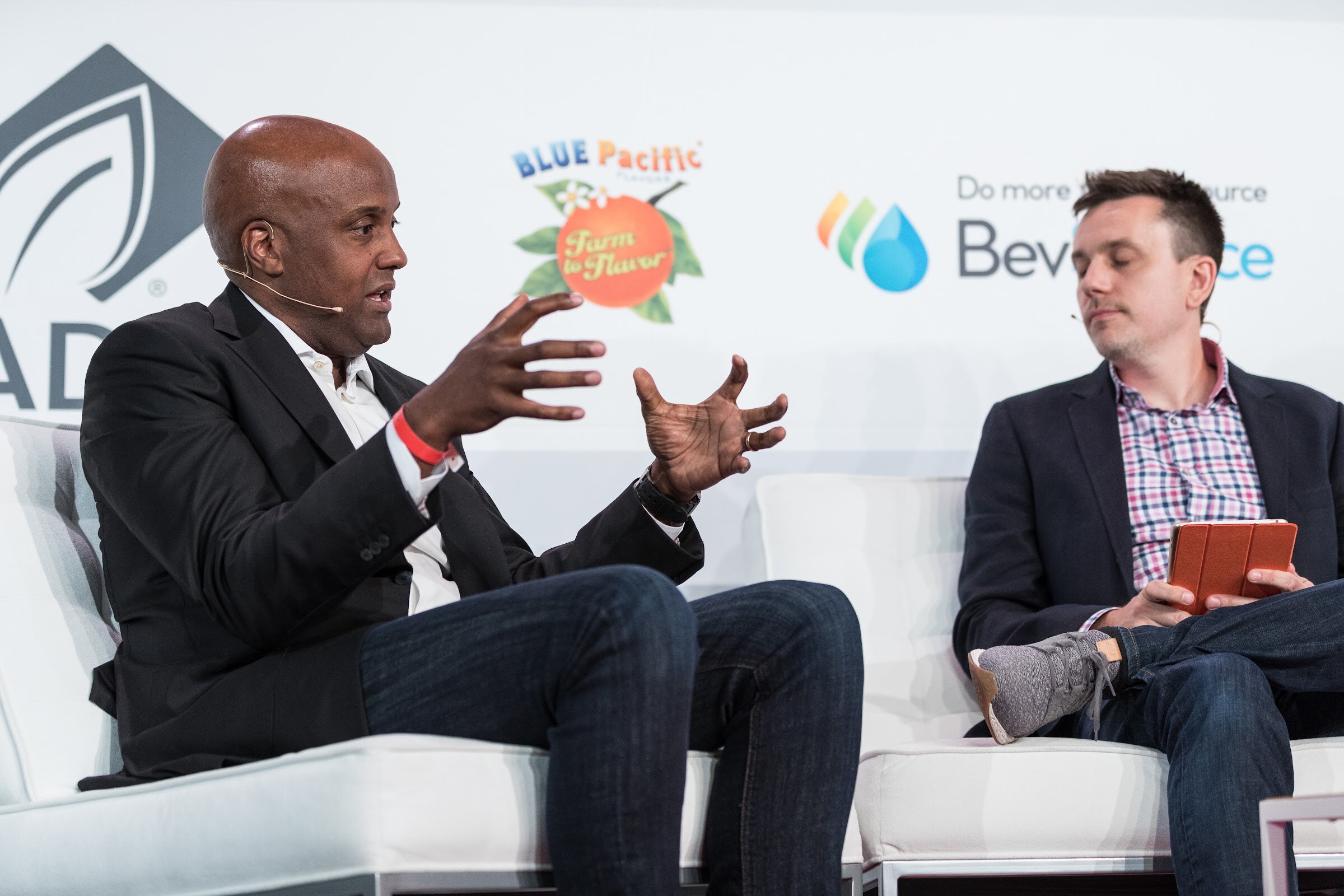 Two men on white chairs on stage; the one at left gestures with both hands while the other holds an orange tablet, in front of ADM, Blue Pacific Flavors and BevSource logos.