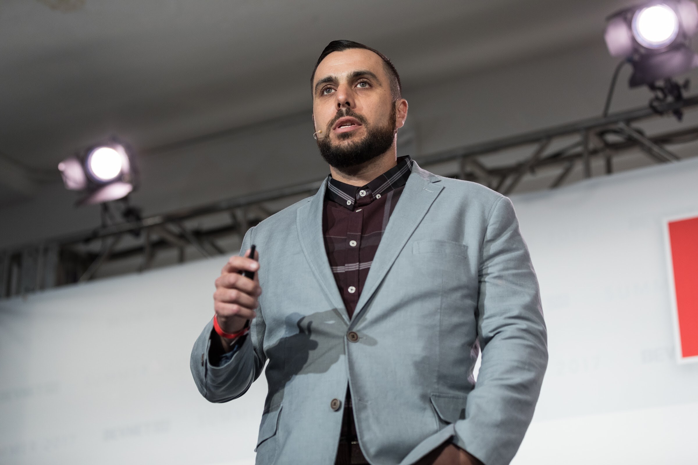Bearded man in a light gray blazer over a dark plaid shirt, holding a clicker, under stage lights.